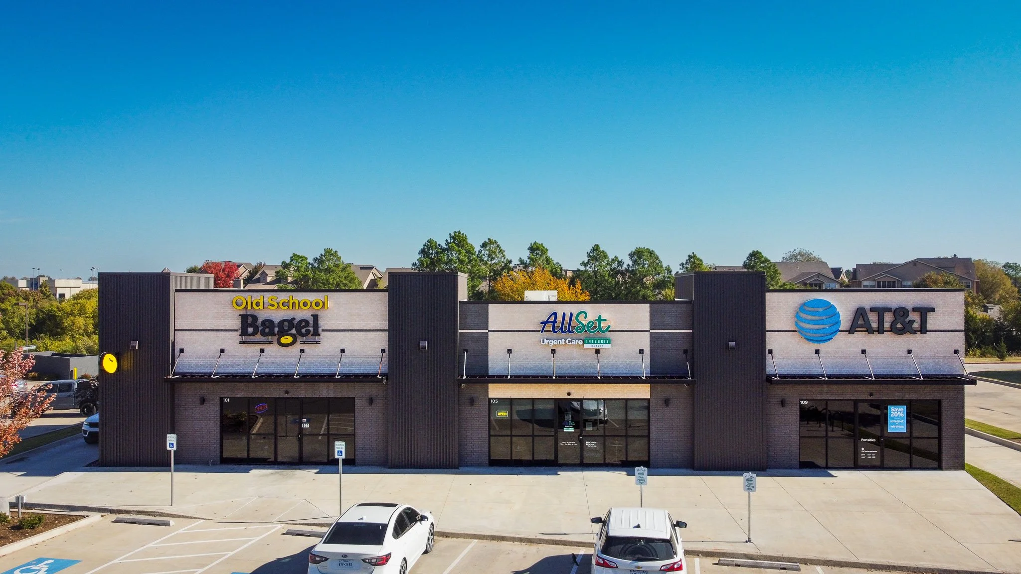 A strip mall with signs for Old School Bagel, All Set Urgent Care, and AT&T, with parking lot in front and trees and houses in the background under a clear blue sky.
