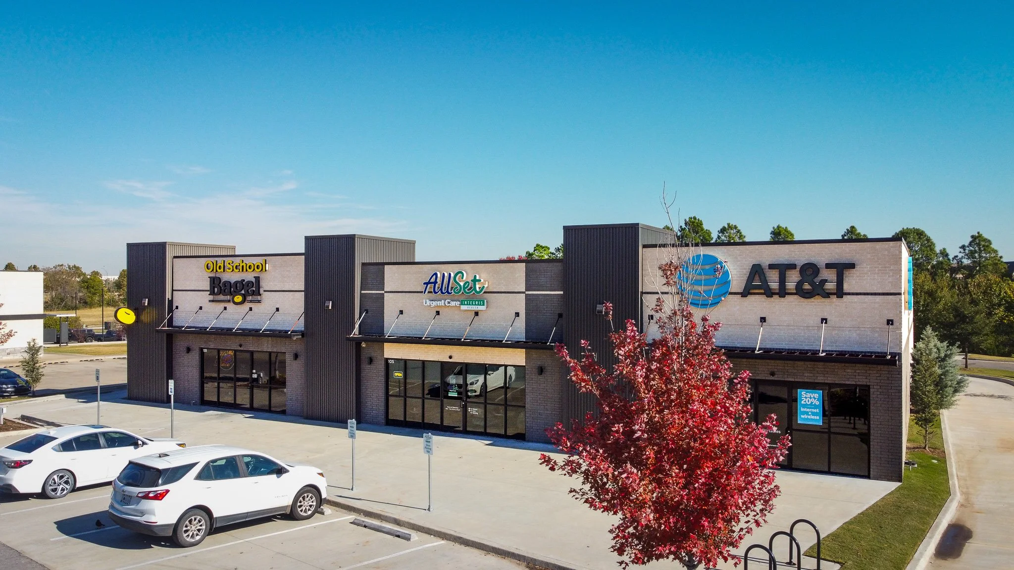 Shopping plaza featuring stores including Old School Bagel, AllSet Urgent Care, and AT&T, with parked cars and a red-leafed tree in the foreground.