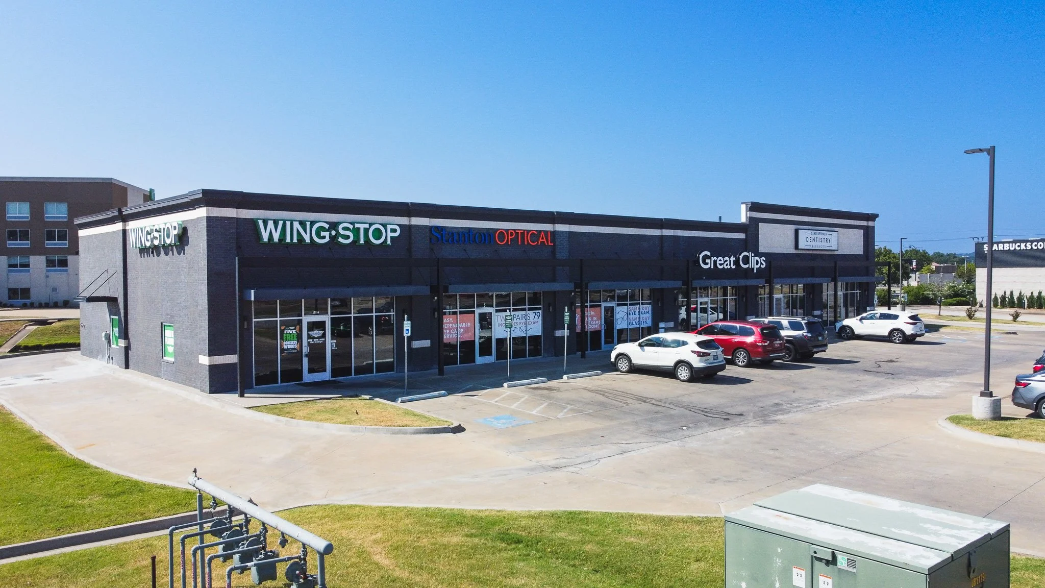 A parking lot in front of a strip mall with signs for Wing Stop, Stanton Optical, Great Clips, and other businesses under a clear blue sky.