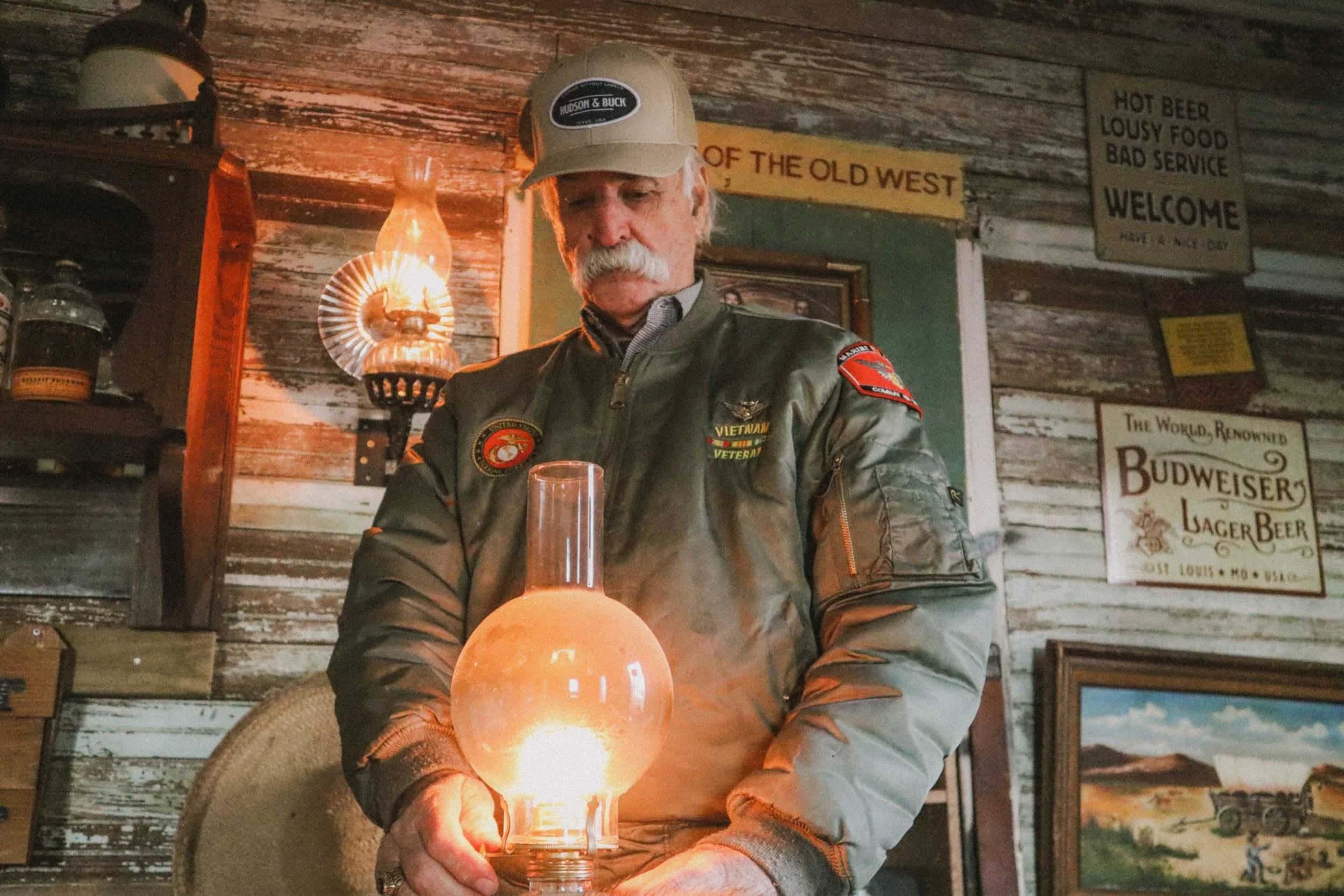 A man with a gray mustache wearing a beige cap, gray jacket, and patches on the sleeves is sitting in a rustic wooden room, holding a glowing orange glass object, with vintage signs and pictures on the wall behind him.