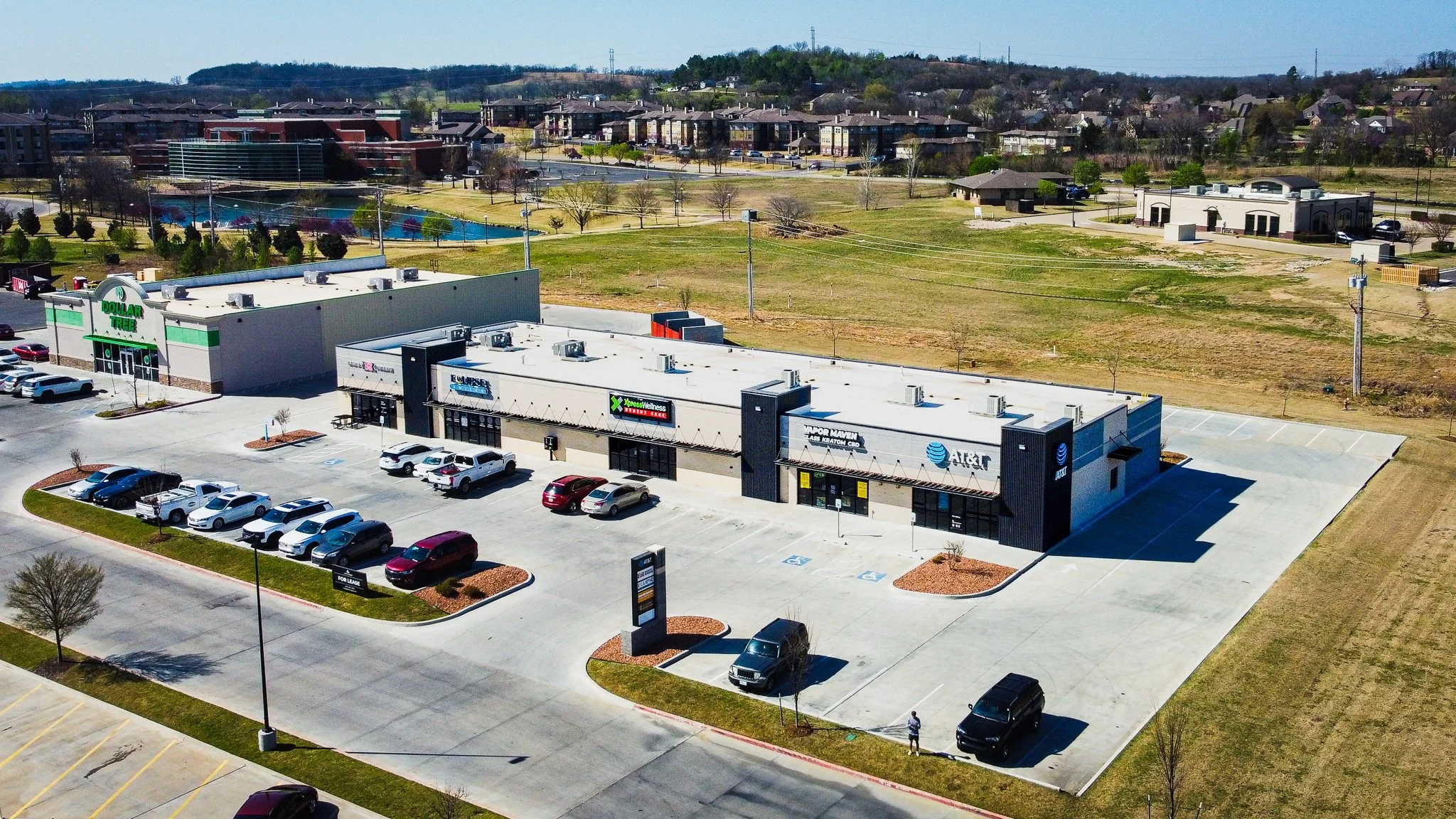 Aerial view of a shopping center with parking lot, surrounded by residential buildings and green spaces.