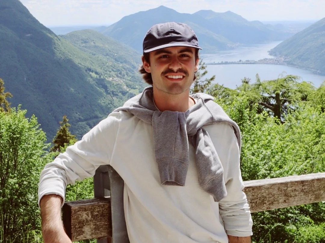 A smiling young man with a gray hoodie draped over his shoulders, wearing a gray cap, standing outdoors on a wooden railing with a mountain and lake landscape in the background.