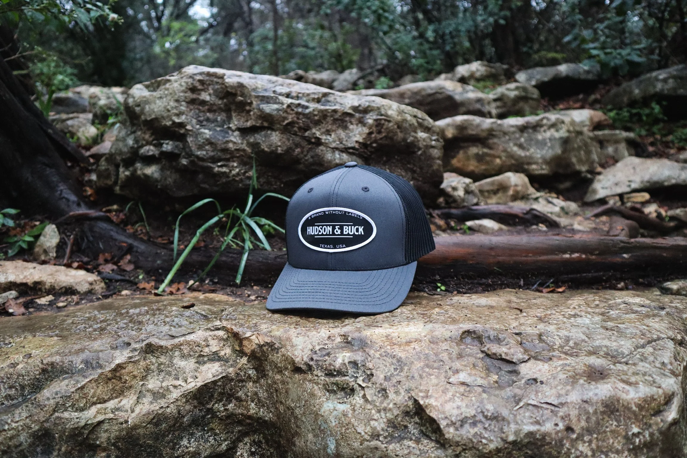 A gray trucker hat with a black mesh back and a white logo patch reading "Hudson & Buck" resting on a rock in a forest setting with large rocks and trees in the background.