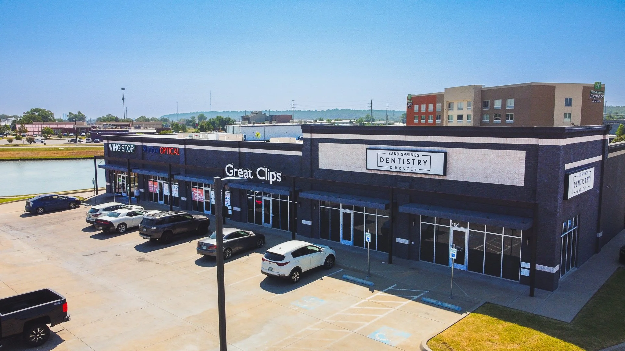 A commercial shopping complex with various stores, parking lot with cars, and surrounding buildings under a clear blue sky.