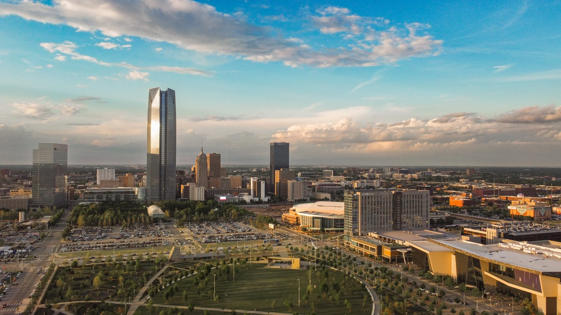 OKC skyline featuring tall skyscrapers, a park with trees and pathways, and a parking lot in the foreground. The sky is partly cloudy with the setting sun reflecting on the buildings.