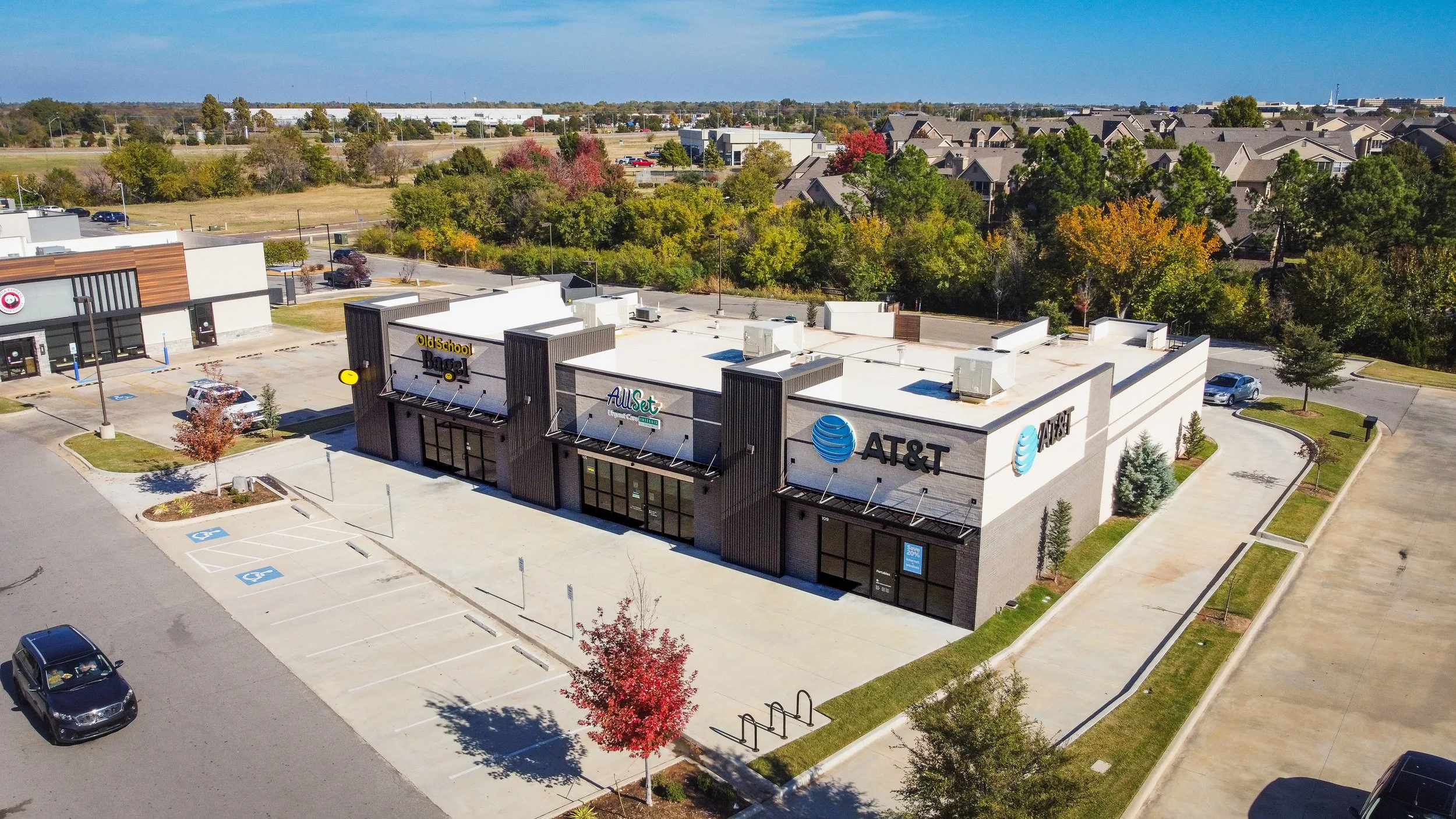 An aerial view of a commercial shopping center with stores including AT&T, Old School Bagel, and All Set. The parking lot has a few cars parked and several empty parking spaces, with accessible parking spots marked. Surrounding the shopping center ar