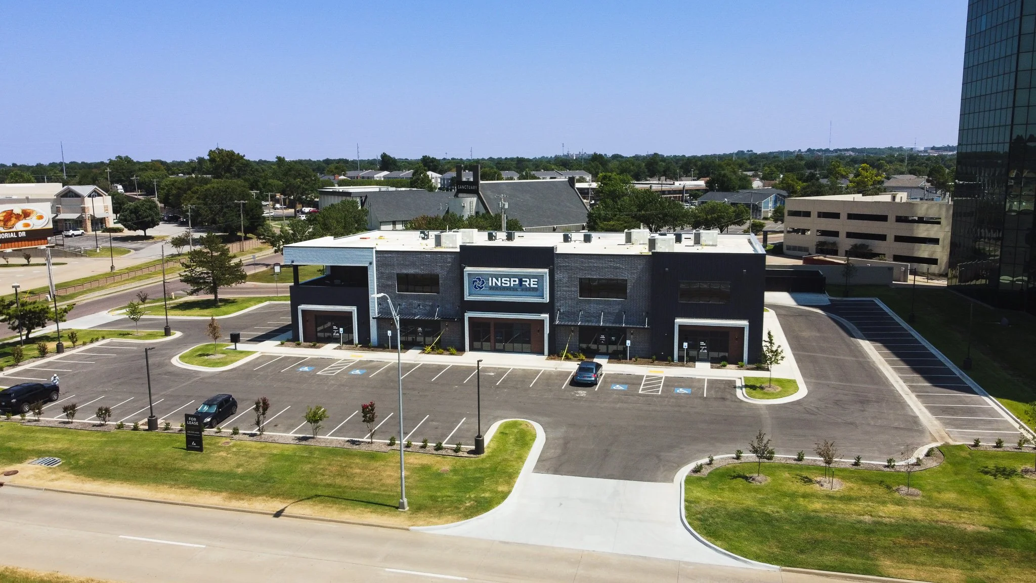 An aerial view of a commercial building with the sign 'INSPIRE' on it, surrounded by parking lots, green trees, and nearby rooftops, with a tall glass office tower on the right side.