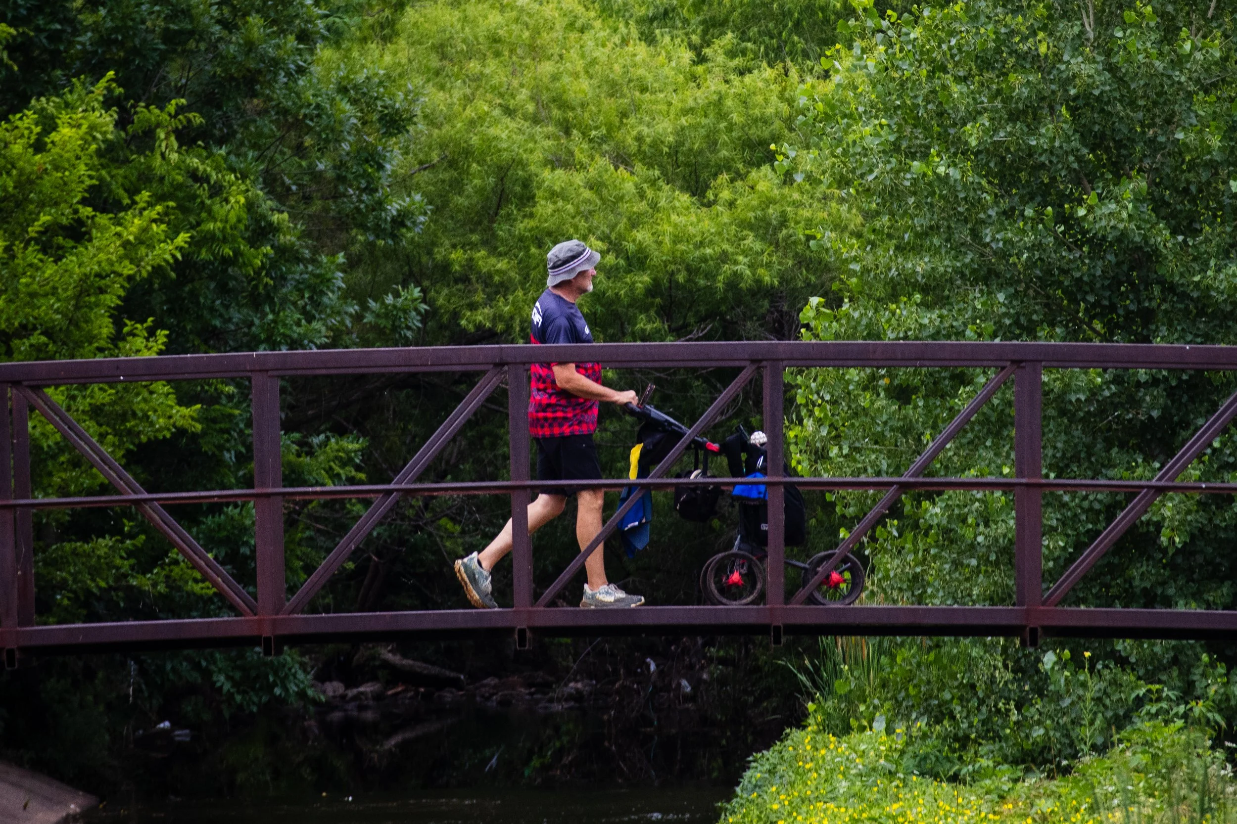 An elderly man wearing a bucket hat and a dark shirt pushes a stroller on a wooden bridge surrounded by lush green trees and vegetation.