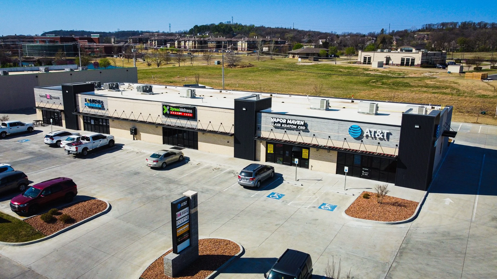 Aerial view of a commercial shopping mall parking lot with several parked cars, including a handicapped parking space, and a building with AT&T and vapor products signage. The background shows a grassy area, trees, and residential buildings under a c