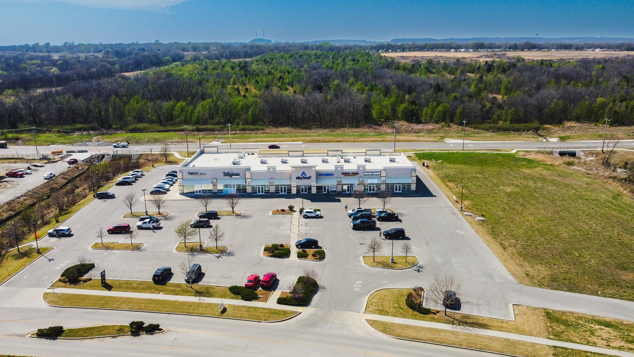 Aerial view of a small shopping plaza with multiple storefronts, parking lot with cars, and surrounding green landscape with trees and open fields.