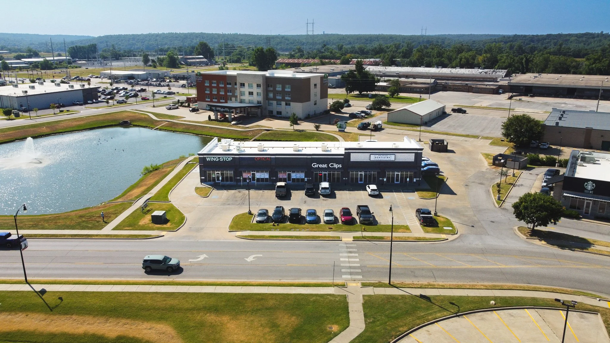 An aerial view of a commercial area with a small pond, parking lots, and various buildings including a Wingstop restaurant, a dental office, and other retail stores, with a backdrop of green hills and transmission towers.
