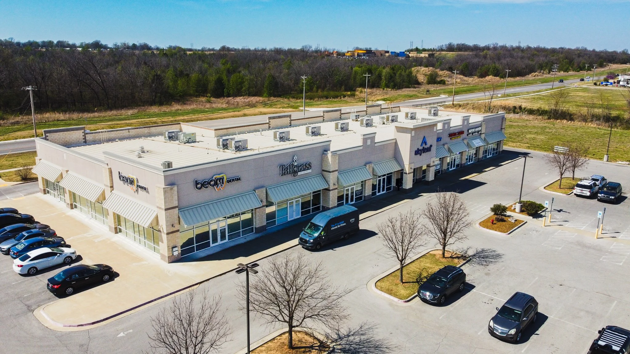 An aerial view of a strip mall with multiple businesses, a parking lot with cars, and a background of trees and open land under a clear blue sky.