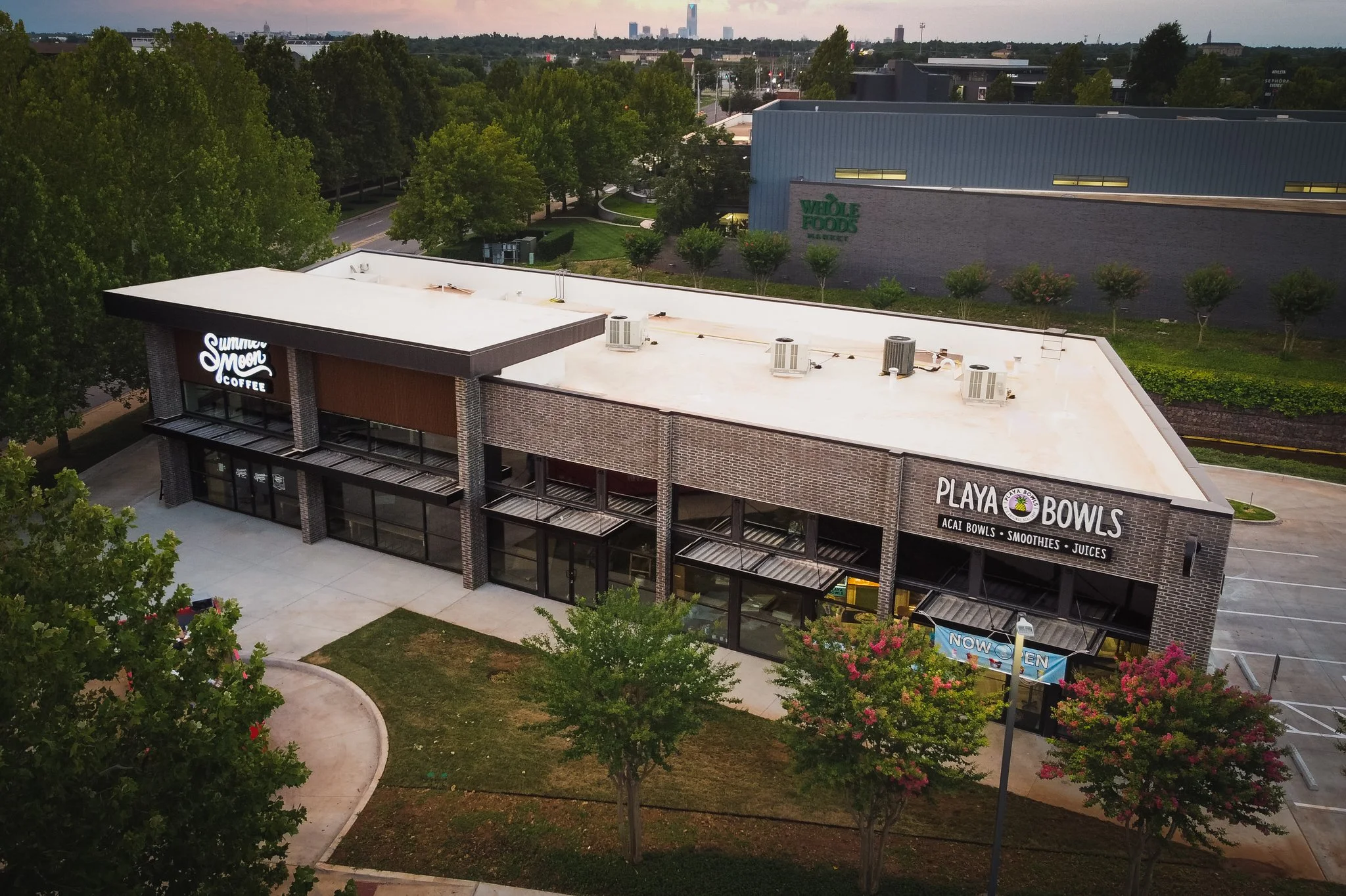 Aerial view of a shopping center with stores including Summer Moon Coffee, Whole Foods Market, and Playa Bowls, surrounded by trees and a parking lot, with a city skyline in the background during the evening.