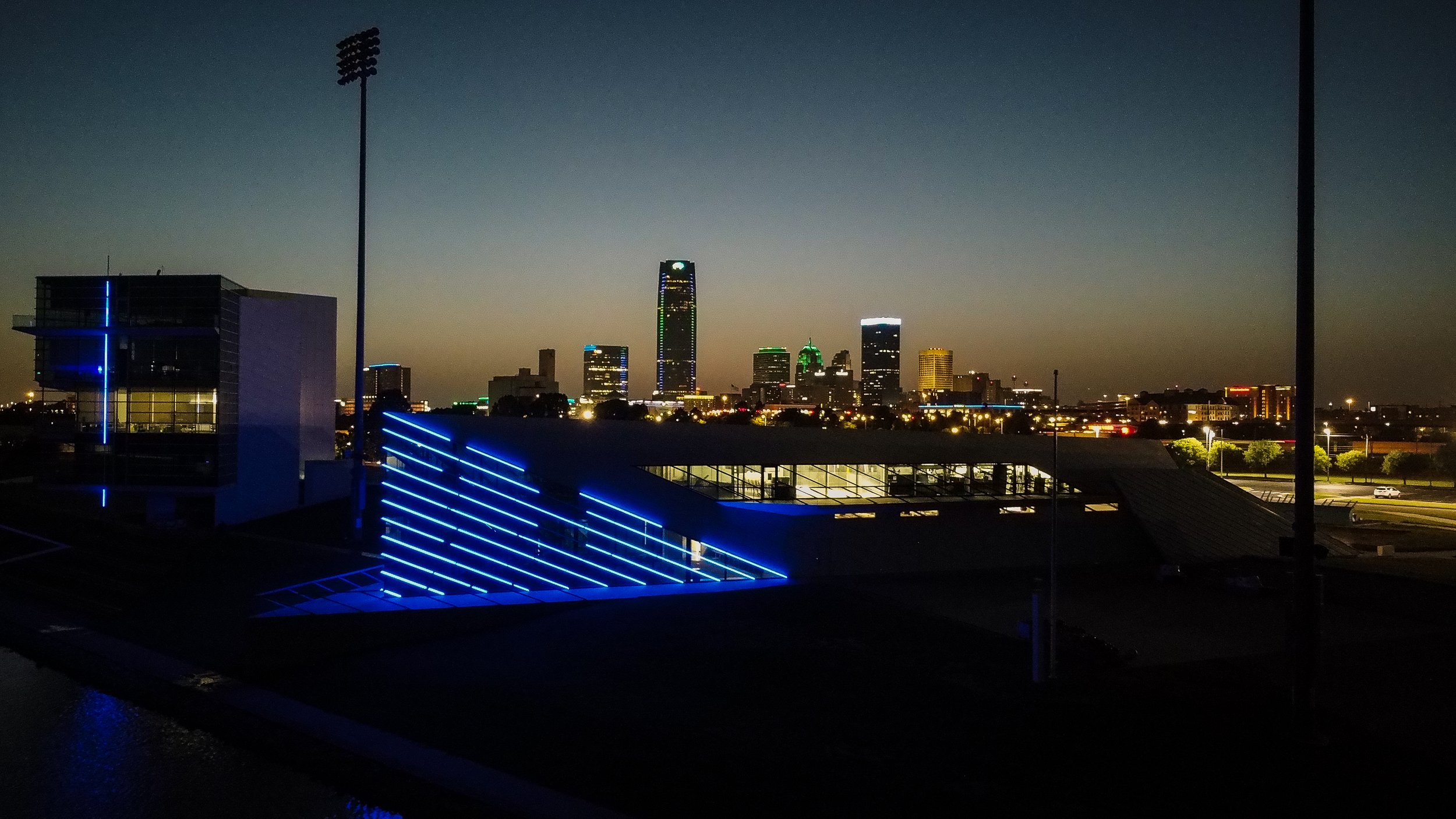 Nighttime city skyline with illuminated skyscrapers, some with green and white lights, and a modern building with blue neon lights in the foreground.