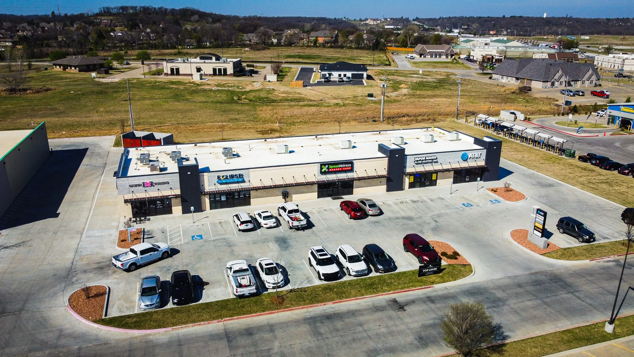 An aerial view of a strip mall with several storefronts, a parking lot with cars, and a grassy field and residential area in the background. The storefronts include businesses like a vape shop, wellness clinic, and a nail salon, with signs visible. T