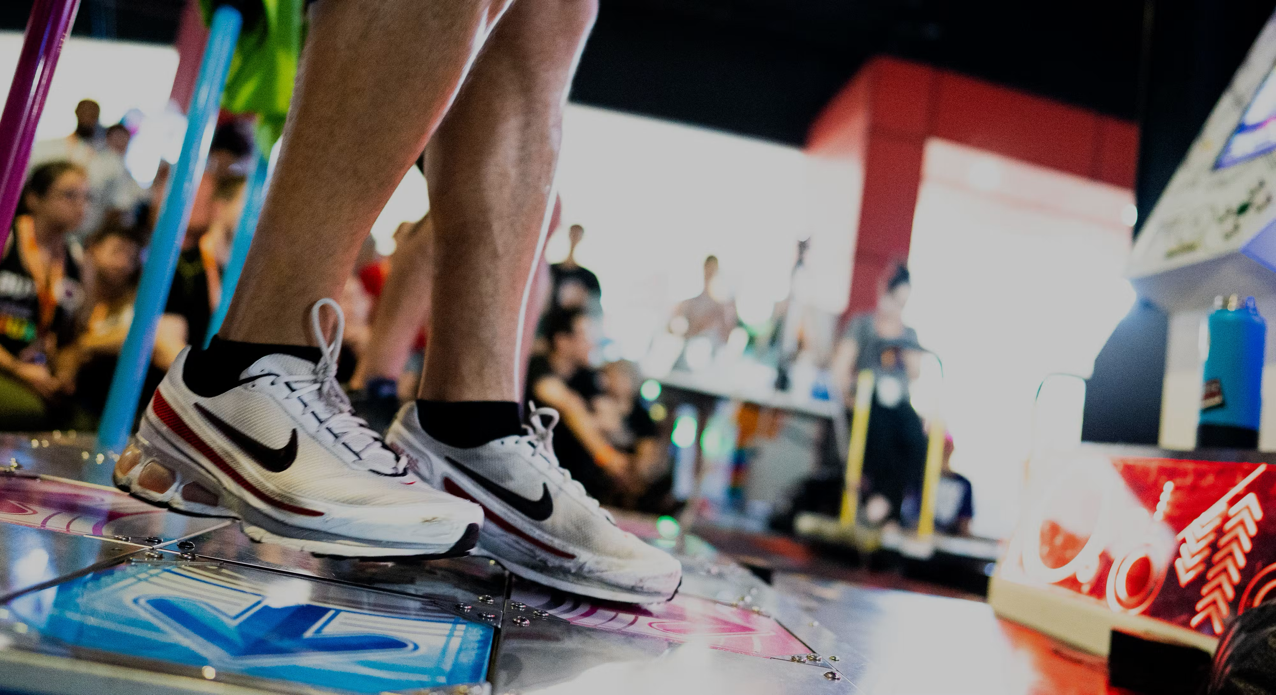 Close-up of person's legs and white Nike sneakers standing on a skateboarding machine or platform at an indoor event with a crowd in the background.