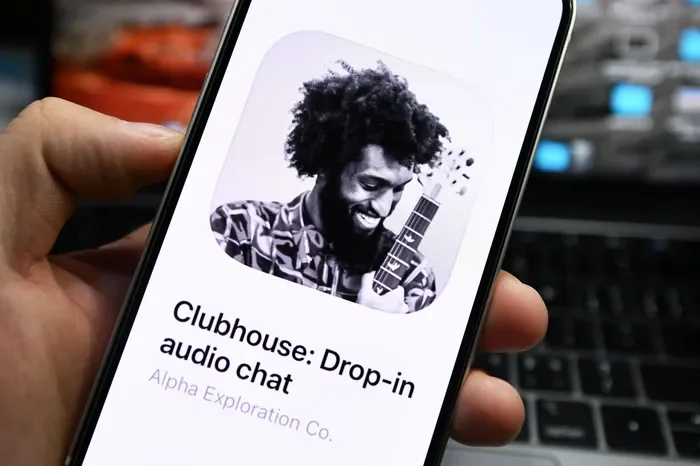 Smartphone screen showing a profile picture of a man with curly hair and a beard, smiling and holding a guitar, with text that reads 'Clubhouse: Drop-in audio chat' and 'Alpha Exploration Co.'
