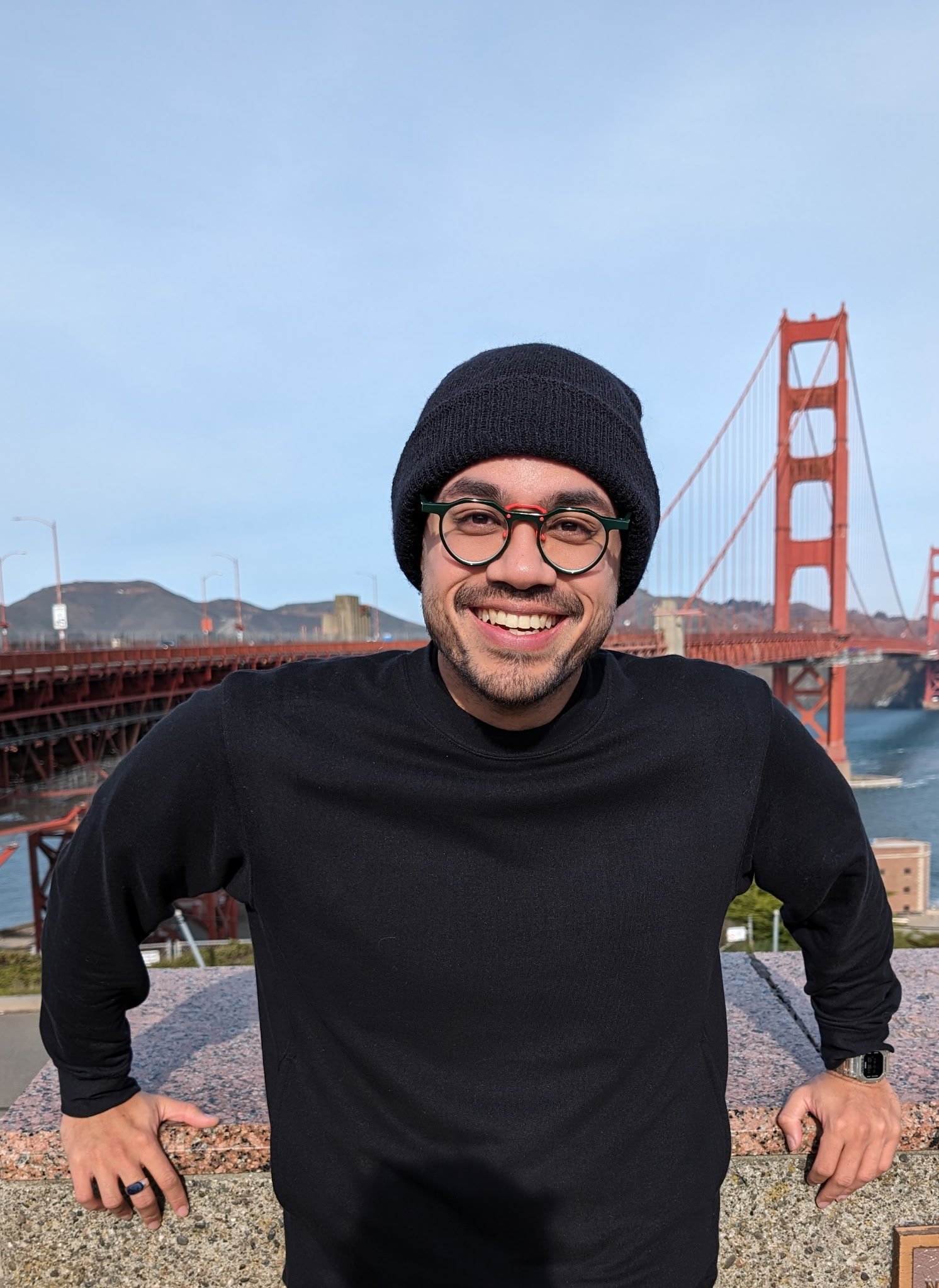 A smiling man wearing glasses, a black beanie, and a black shirt, standing with hands on hips in front of the Golden Gate Bridge in San Francisco.
