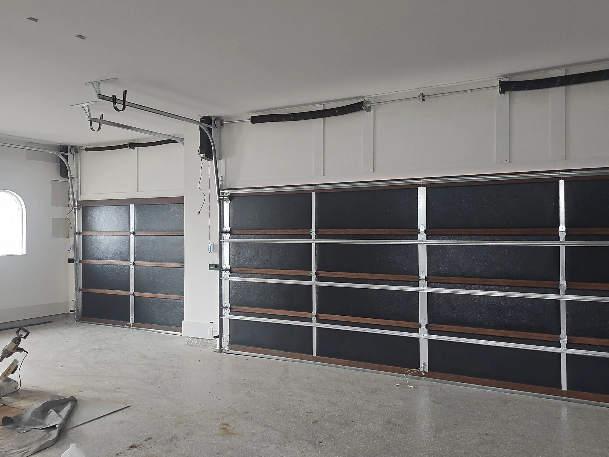 Garage interior with black garage doors, partially installed wall panels, and tools on the unfinished concrete floor.