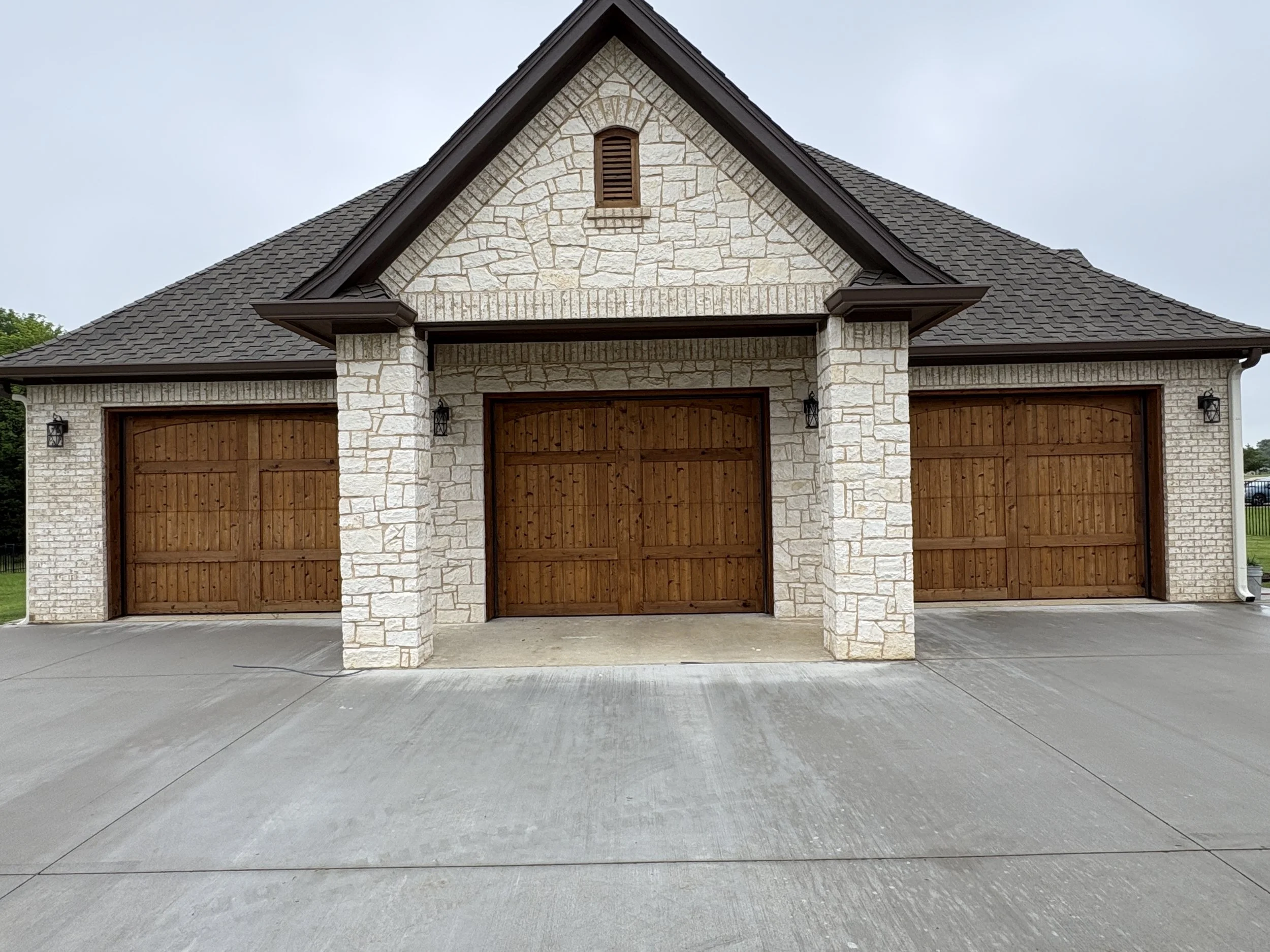 Front view of a house with stone walls, wooden garage doors, and a gabled roof, with a concrete driveway in front.