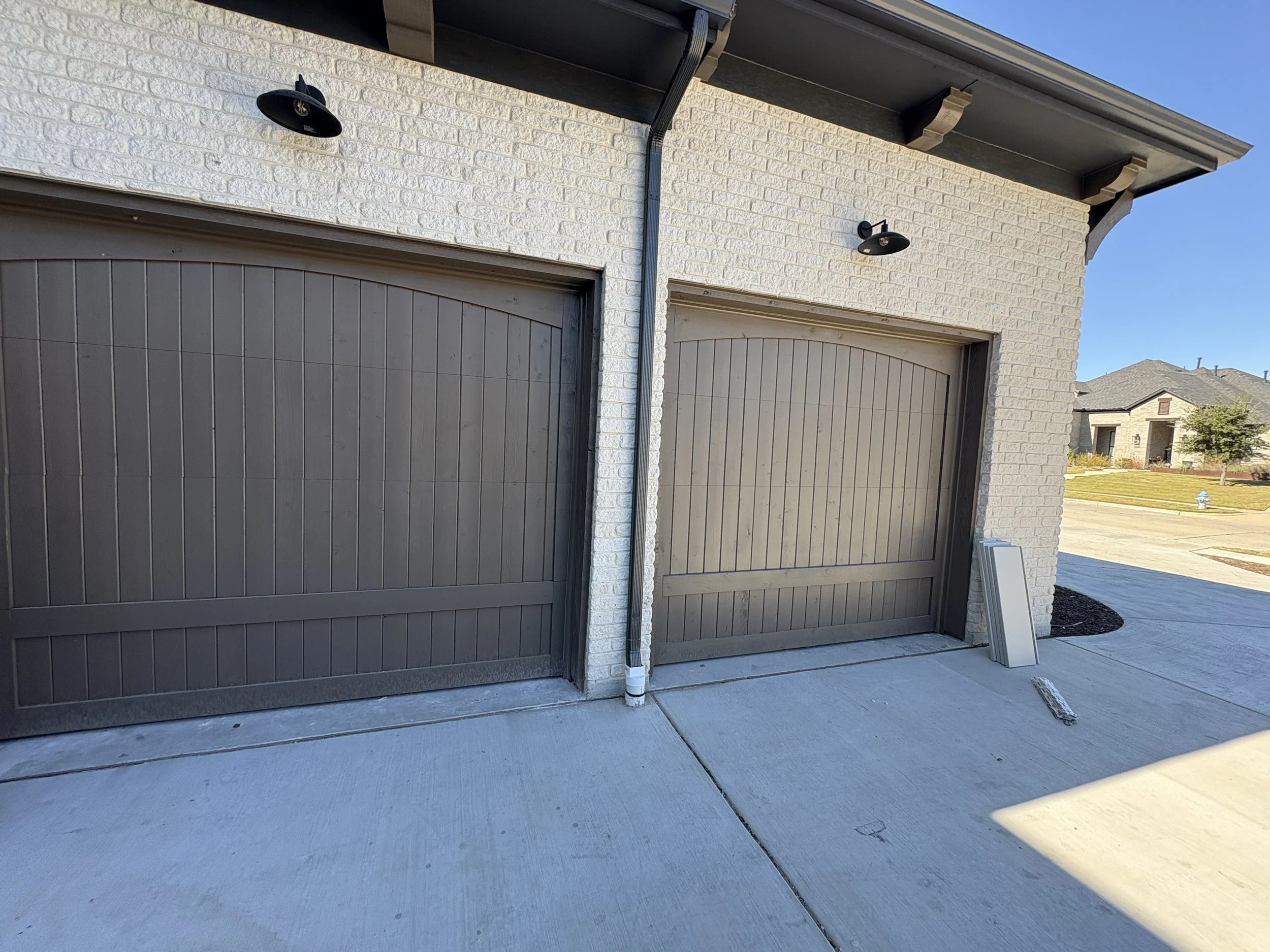 View of garage doors and brick wall of a house with exterior lighting fixtures and a gutter system, in a suburban neighborhood on a sunny day.