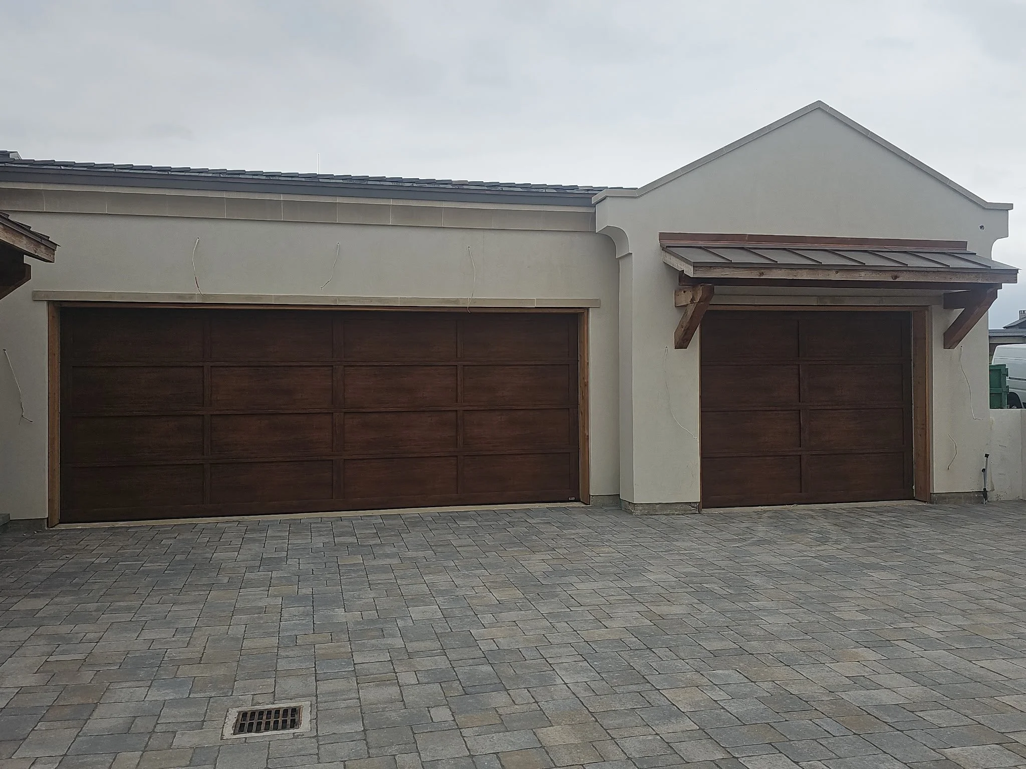 Front view of a house with a brown garage door, beige stucco walls, and a sloped roof with dark shingles. The driveway is paved with interlocking bricks, and a drainage grate is visible on the ground. There are some wires hanging from the wall, possi