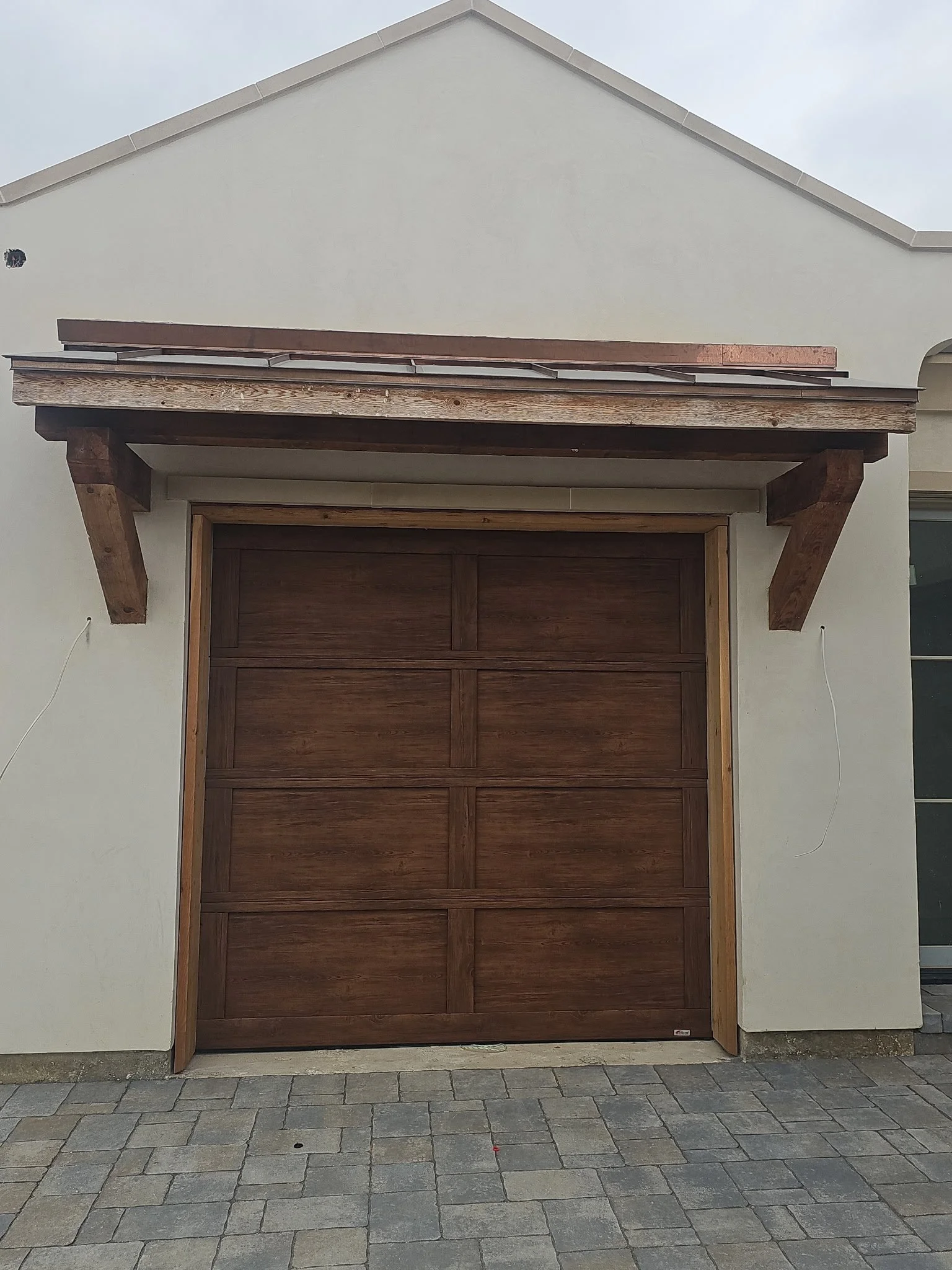 The image shows a closed wooden garage door with a dark brown finish, surrounded by a white exterior wall. Above the garage door, there is a small wooden awning supported by two angled wooden brackets, with a piece of brown material lying on top of t