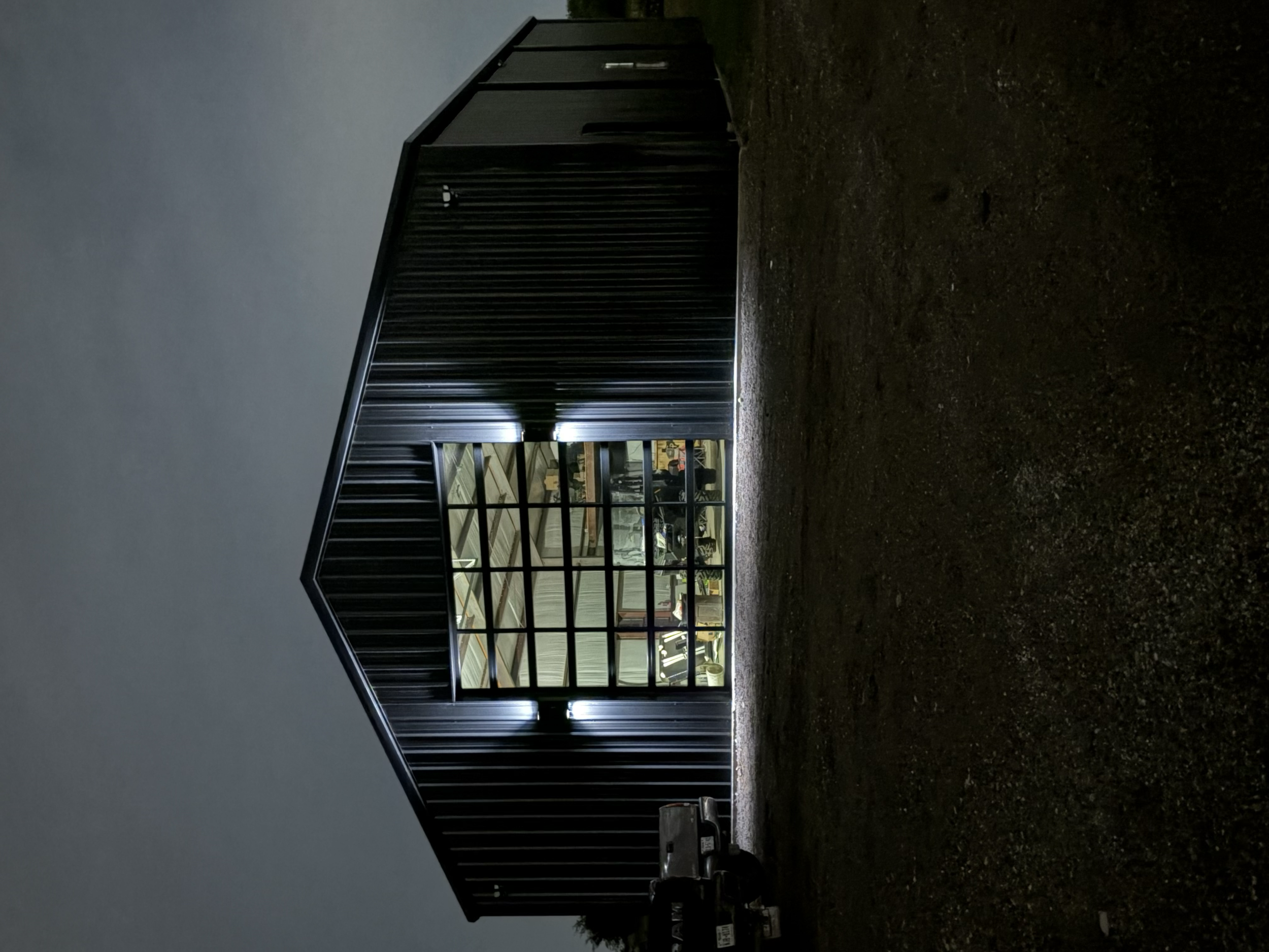 A view of a commercial building's exterior at night with large glass windows revealing the interior, and black metal siding, with a gravel ground in front.