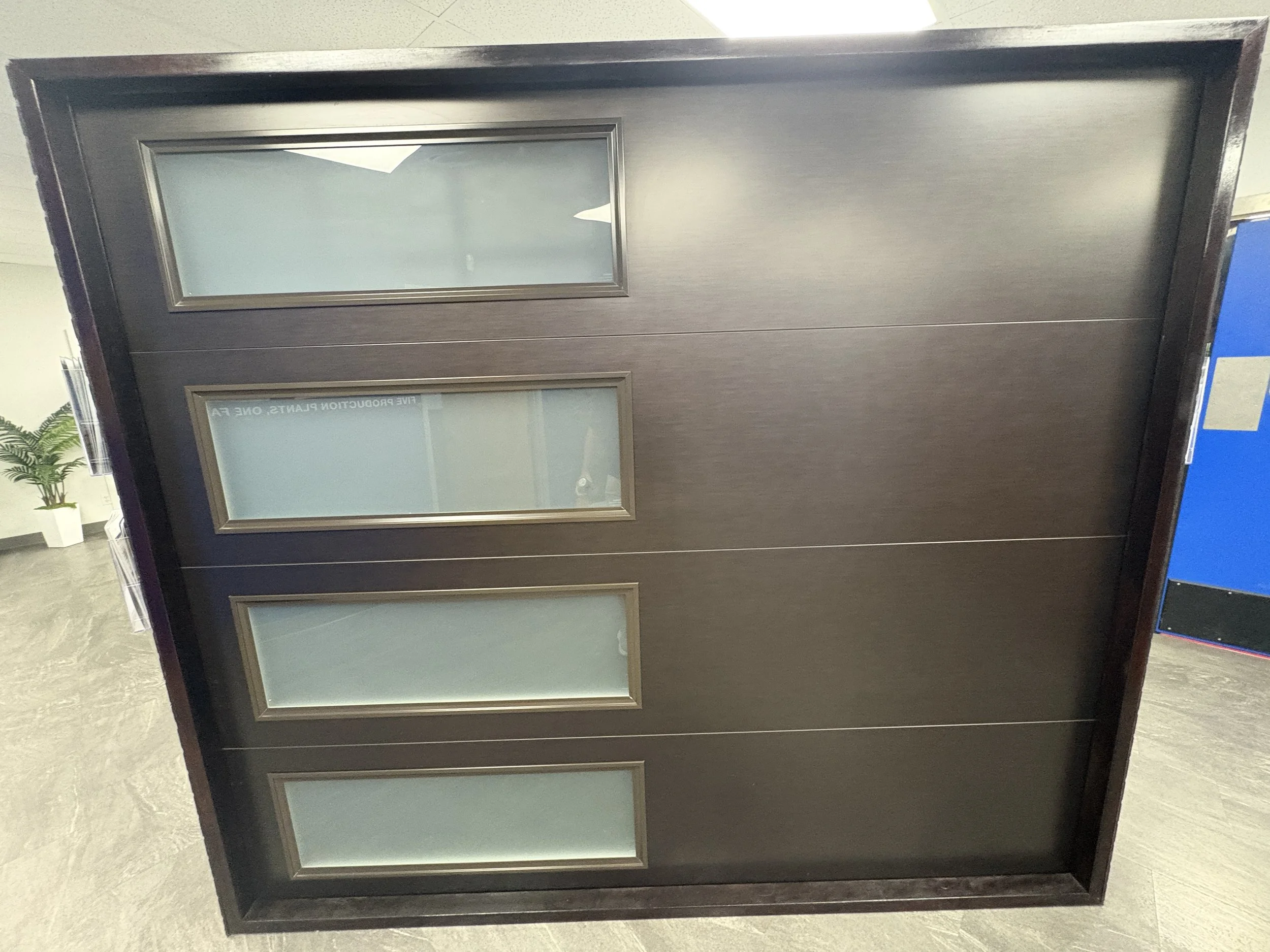 Dark-colored door with three rectangular frosted glass panels in an indoor setting, with a small potted plant and blue object partially visible on the side.
