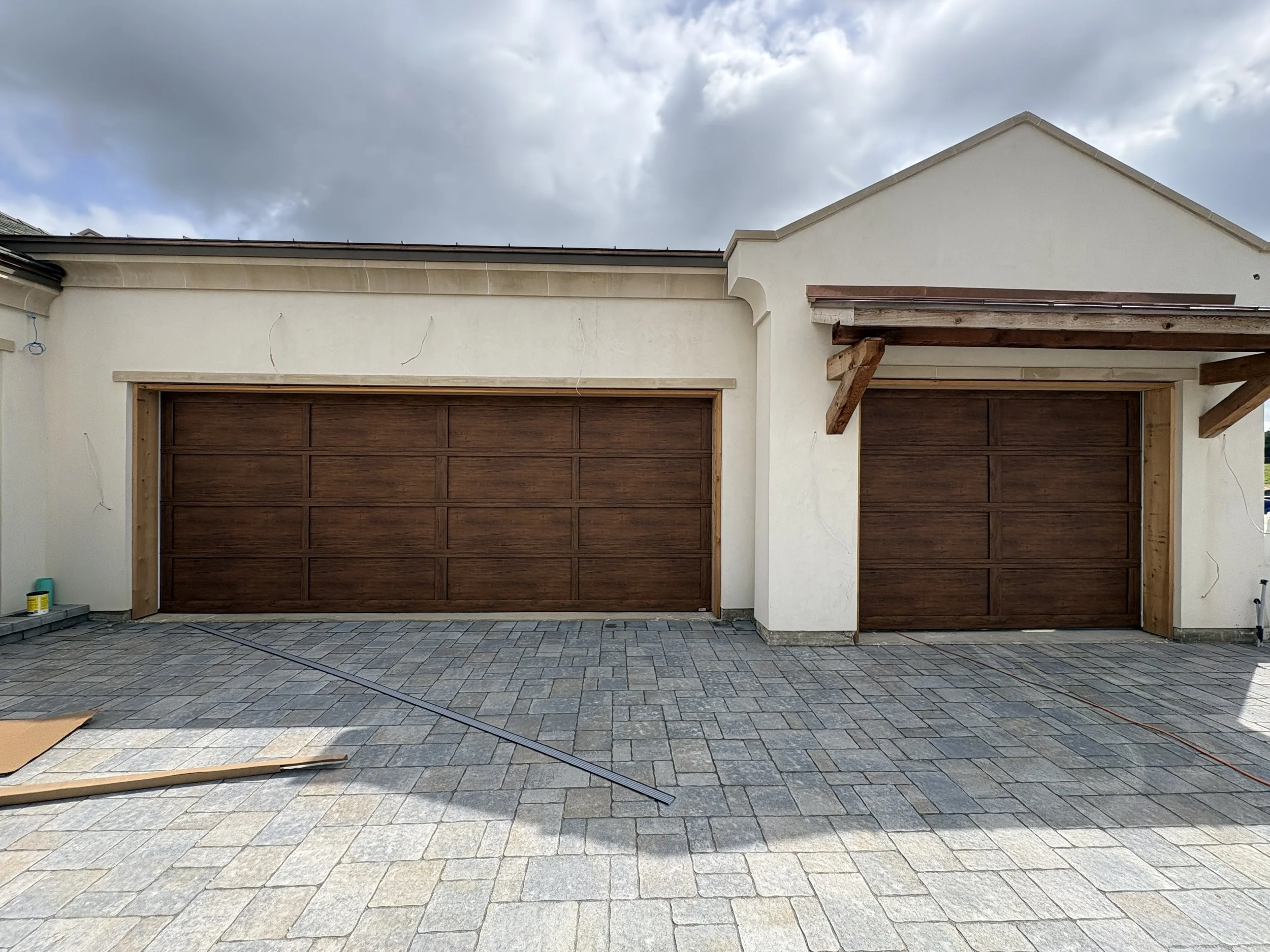 The image shows a house under construction with two garage doors, a paved driveway, construction materials, and tools in the foreground. The house has white walls, wooden garage doors, and a new roof. The sky above is cloudy.