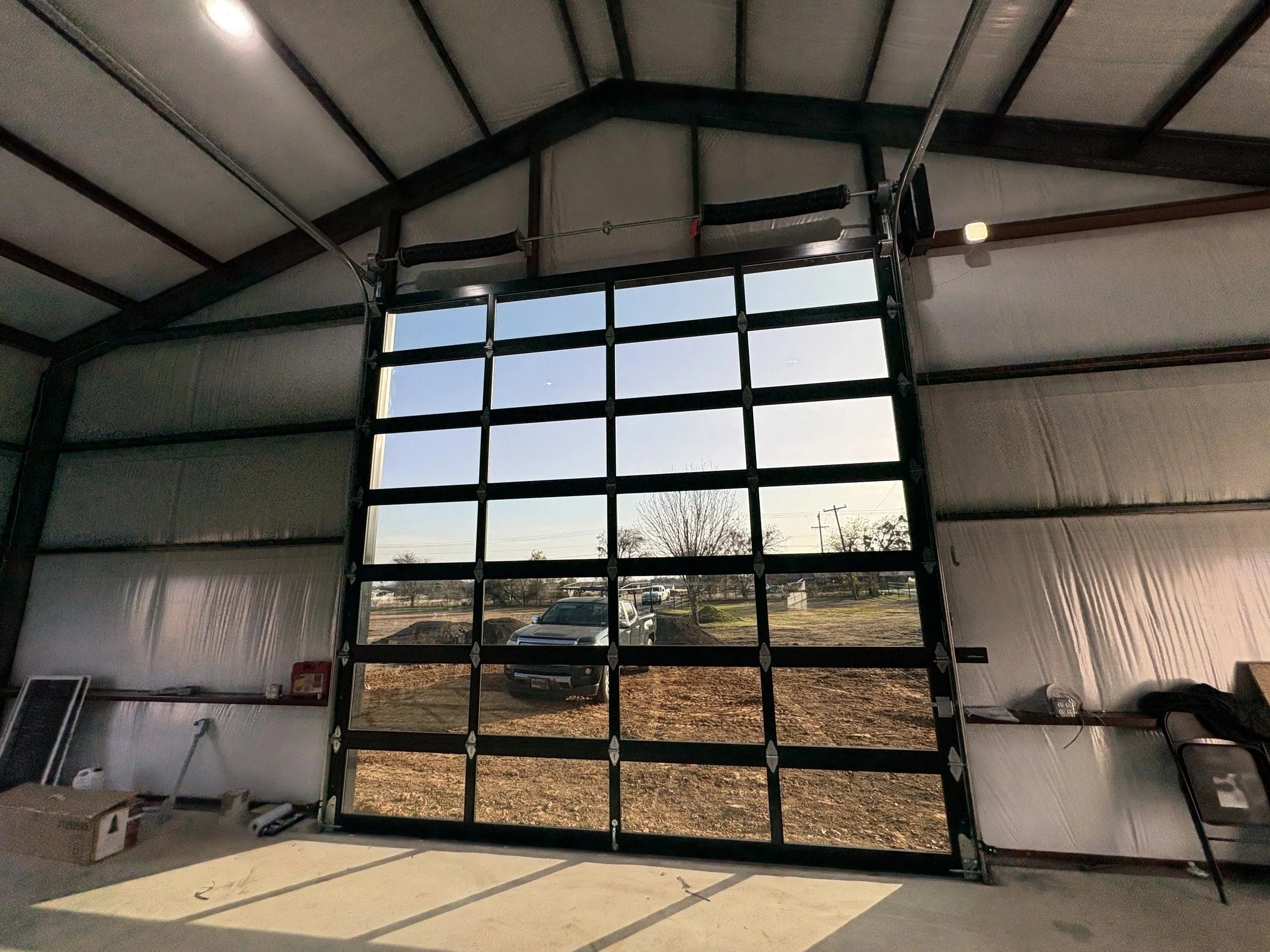 Inside a garage with an open door, showing a parking lot with cars and a tree outside, and some boxes and items inside.