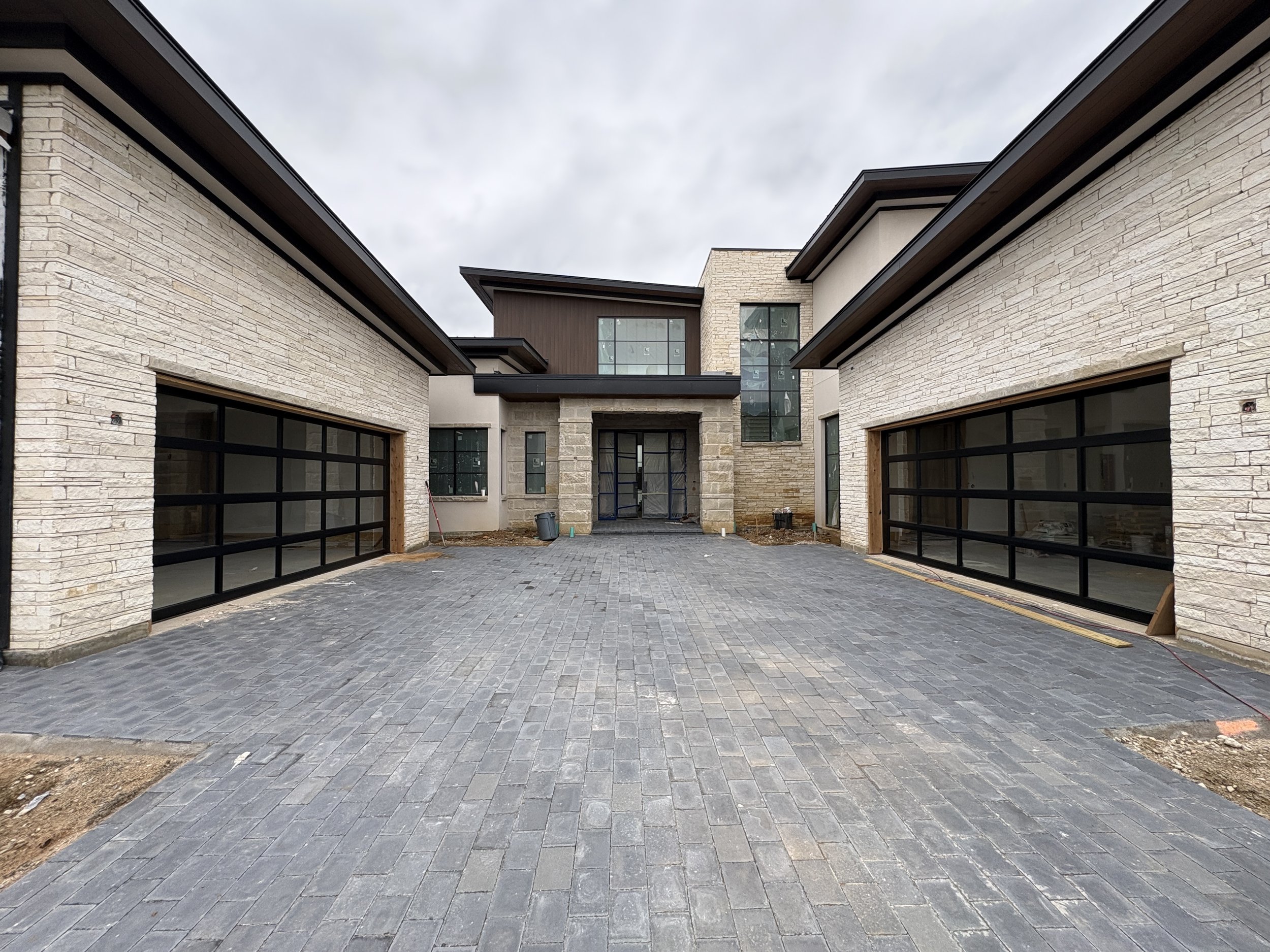 A modern residential building under construction with light-colored brick walls, dark trim, and a paved driveway leading to garage doors. The sky is overcast.