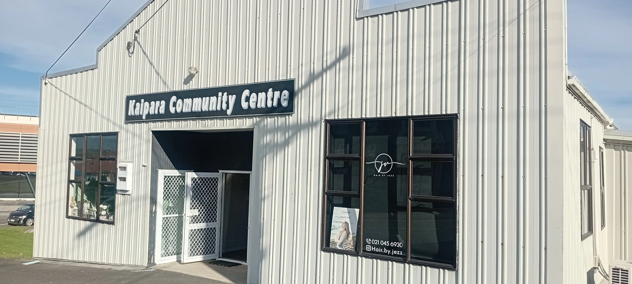 A white, metal-clad building with a sign that reads 'Kaipara Community Centre' on the front. The building has large windows and a small entrance with a gate. There is a sign for a hair salon with a woman’s photo and contact information. The sky is cl