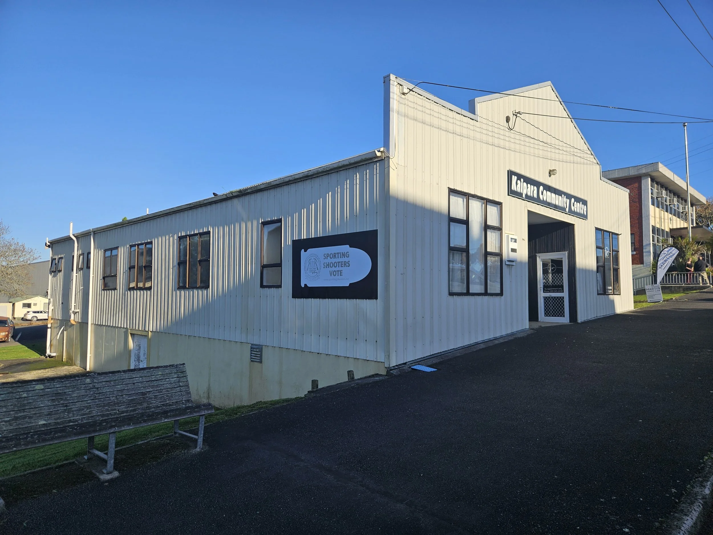 A two-story building labeled 'Kalpara Community Centre' under a bright blue sky. There is a sign on the building that mentions voting and sporting shooters. The building has large windows and a sign with black background and white text. A bench is vi