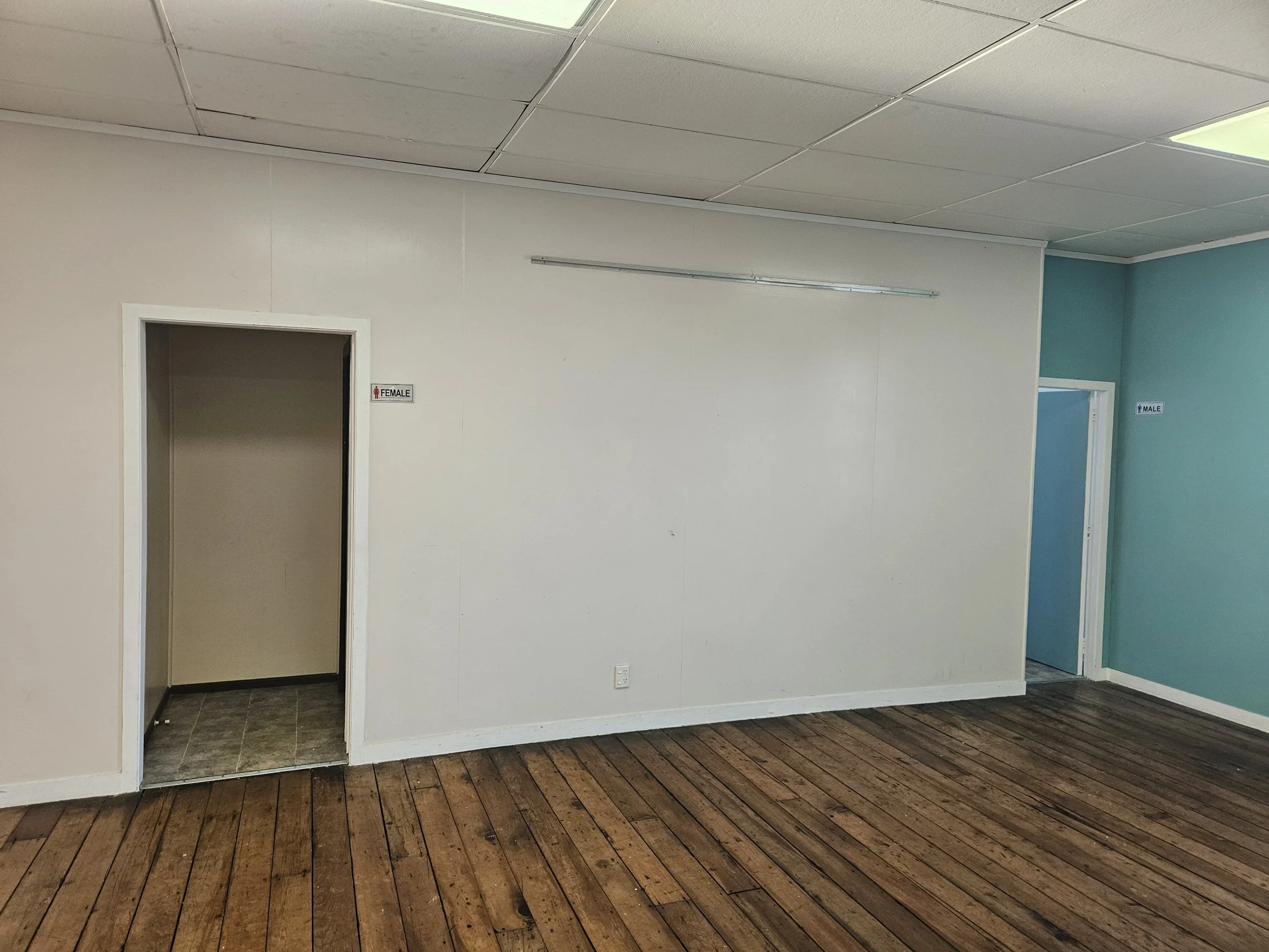 Interior room with white walls and a blue accent wall, wooden flooring, two doorways labeled 'FEMALE' and 'MALE', and a ceiling with suspended tiles and fluorescent lighting.