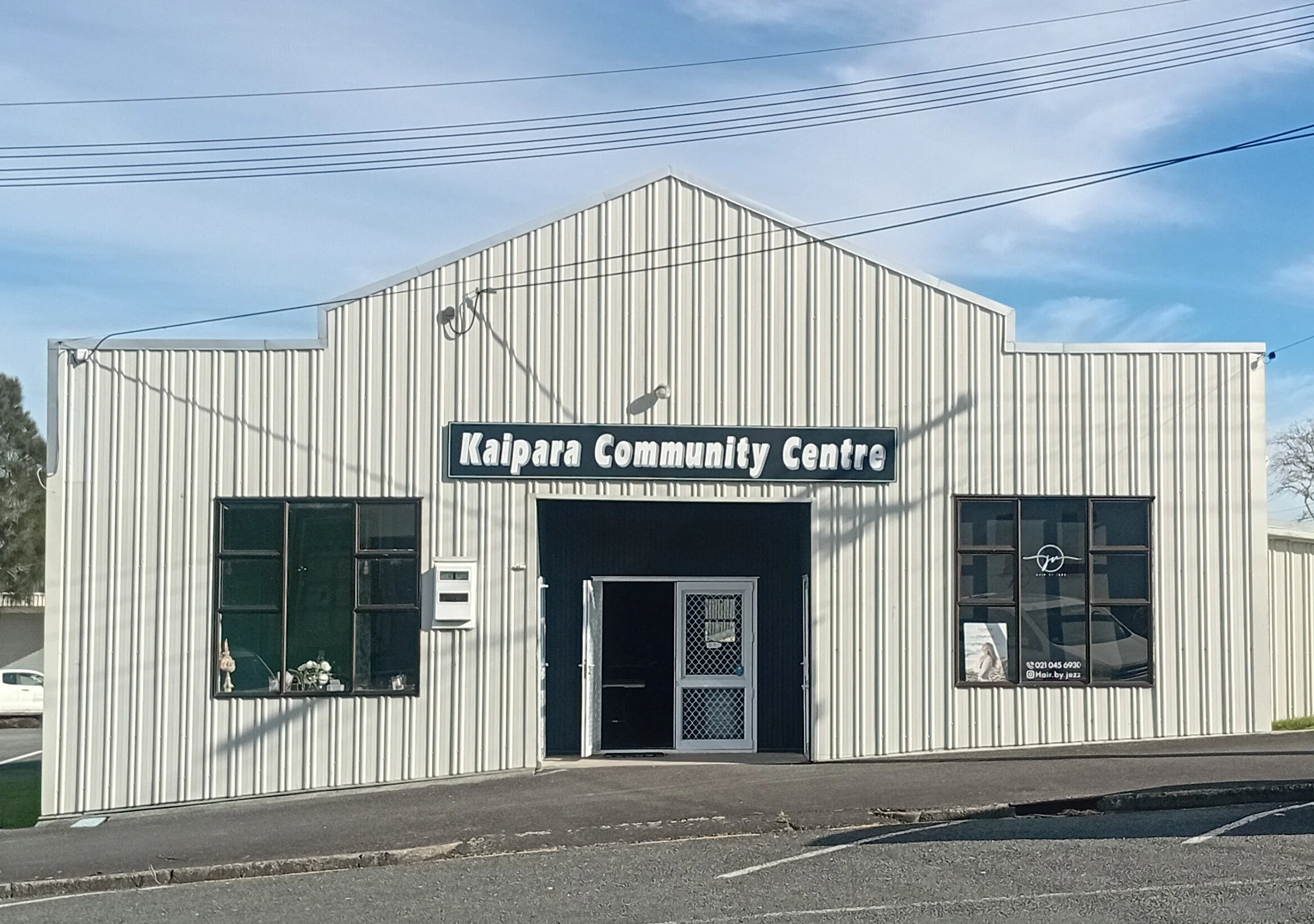 Exterior view of the Kaipara Community Centre building with a metal-sided facade, a sign displaying its name, windows, and a door, under a partly cloudy sky.