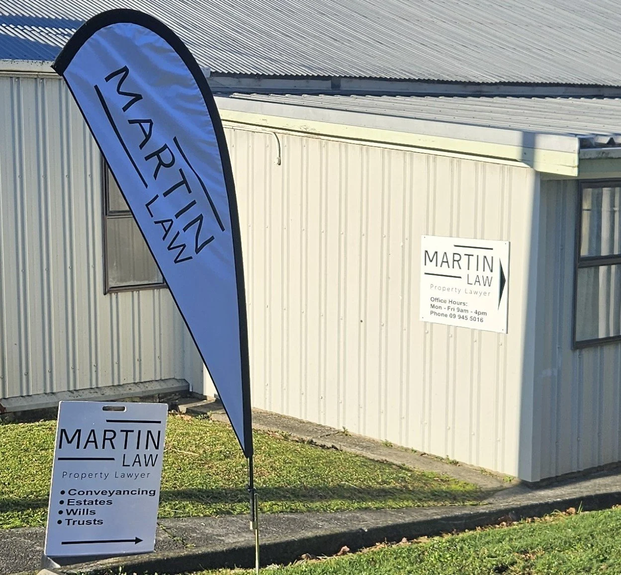 Sign and flag advertising Martin Law, a property lawyer, outside a building with the same signage. The sign lists services like conveyancing, estates, wills, and trusts, with an arrow pointing to the right. The flag displays the Martin Law logo and name.