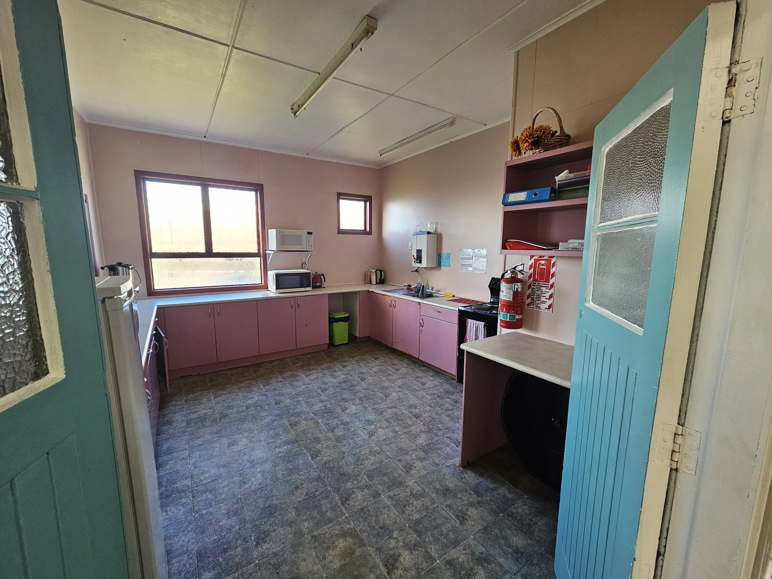 A small kitchen with pink cabinets, a window, a microwave, a fire extinguisher, and a small countertop with some items on it. The photo is taken from outside the room through a door with a blue frame.