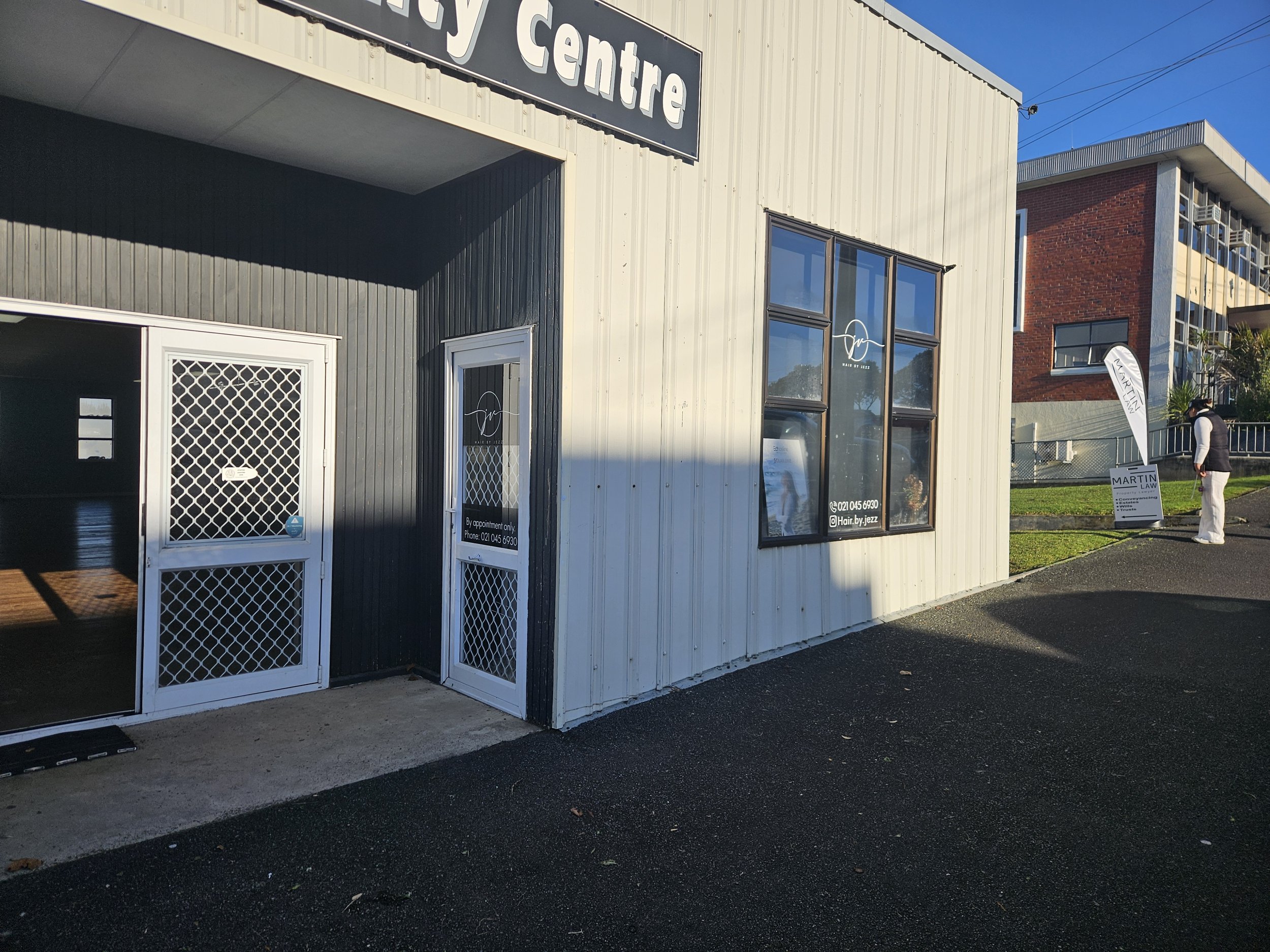 Exterior of a modern building with a sign that reads 'Beauty Centre' at the top. The building has large windows and a sign on the door indicating appointments only. There is a person standing outside next to a sign for Martin Law. The sky is clear an