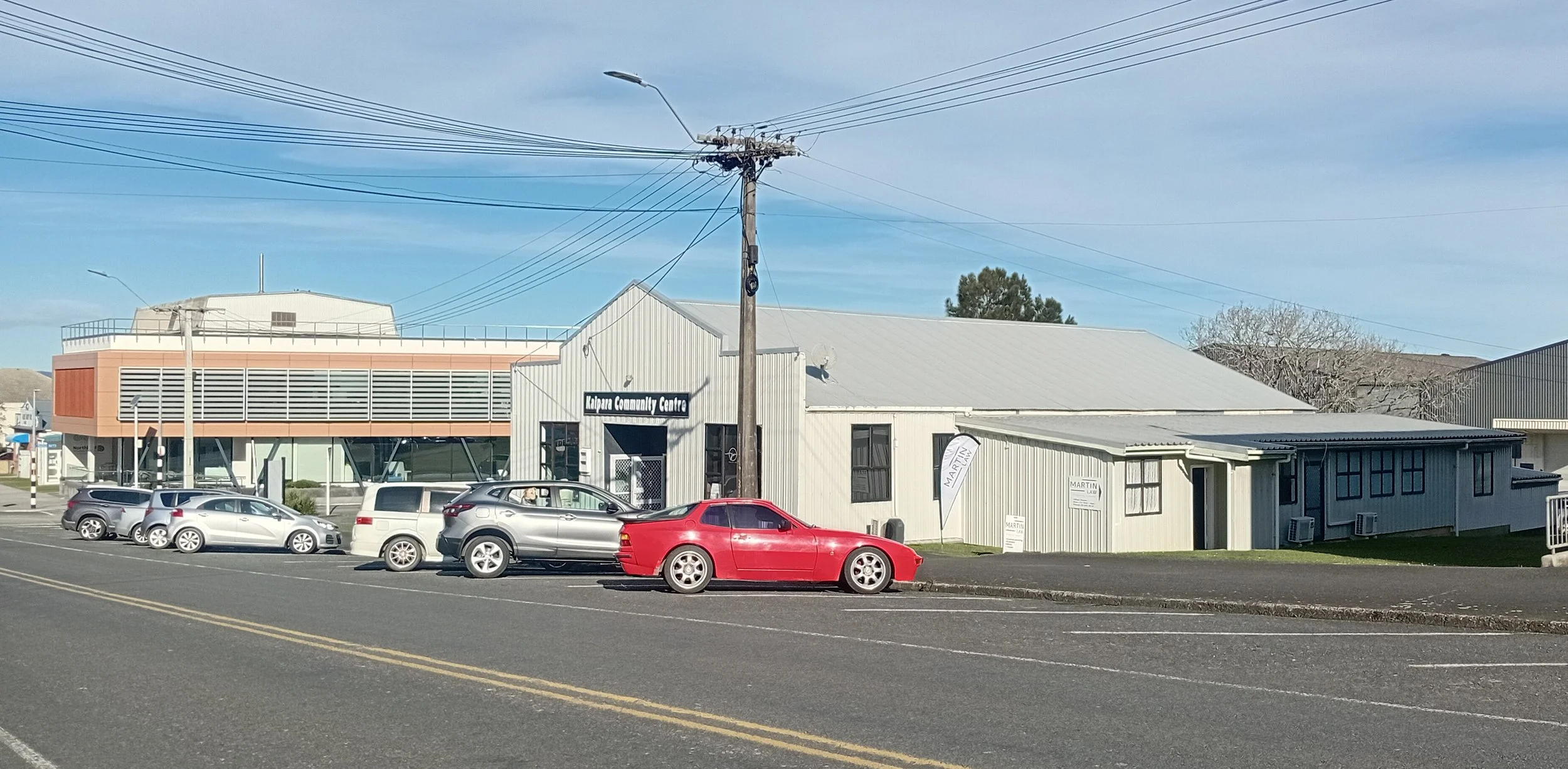 Street view of the Kalipara Community Centre building with parked cars in front and overhead power lines against a blue sky.
