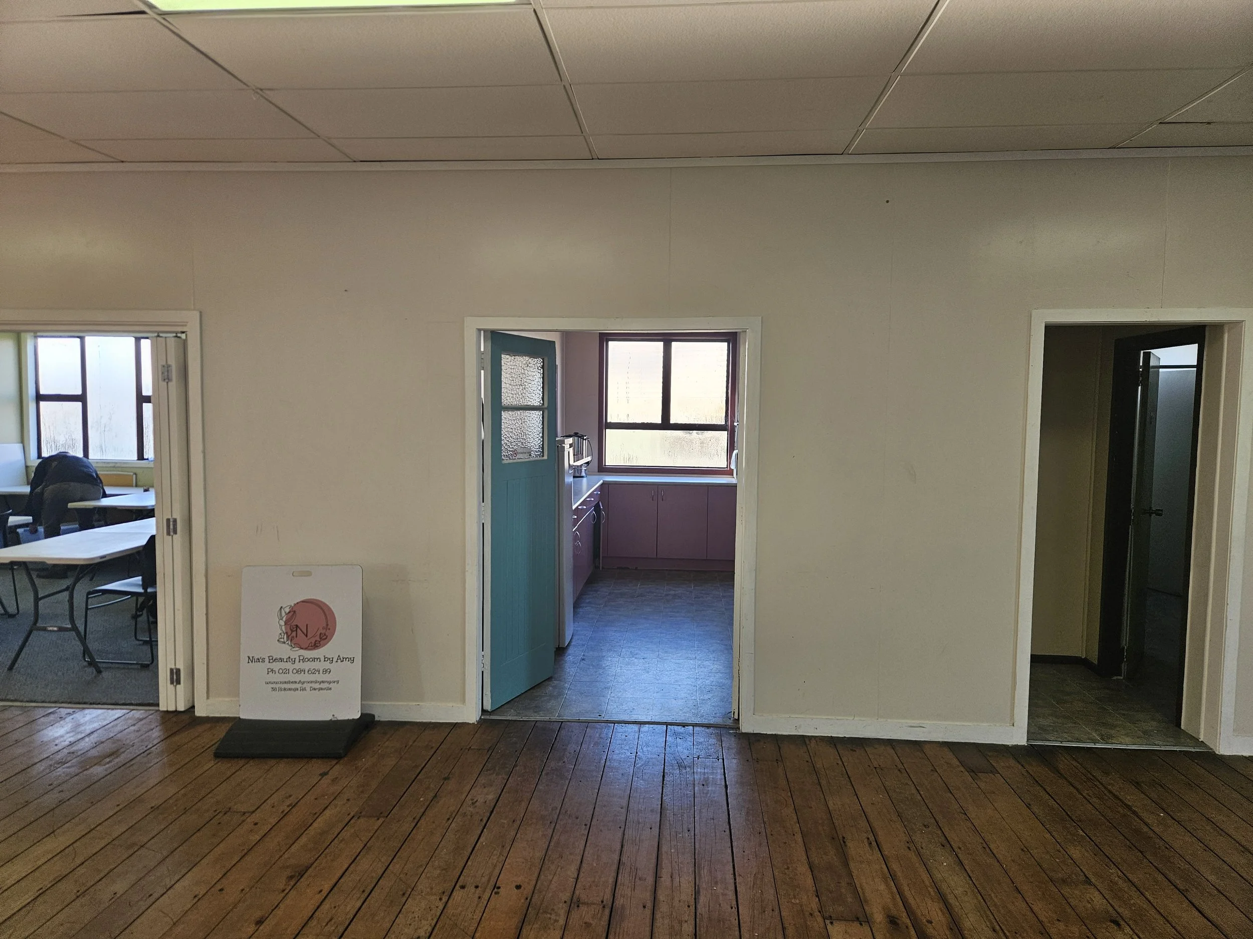 Interior view of a room with a door leading to a kitchen area, seen through framed openings. The room has wood flooring, and a sign for a beauty room is visible on the left side.