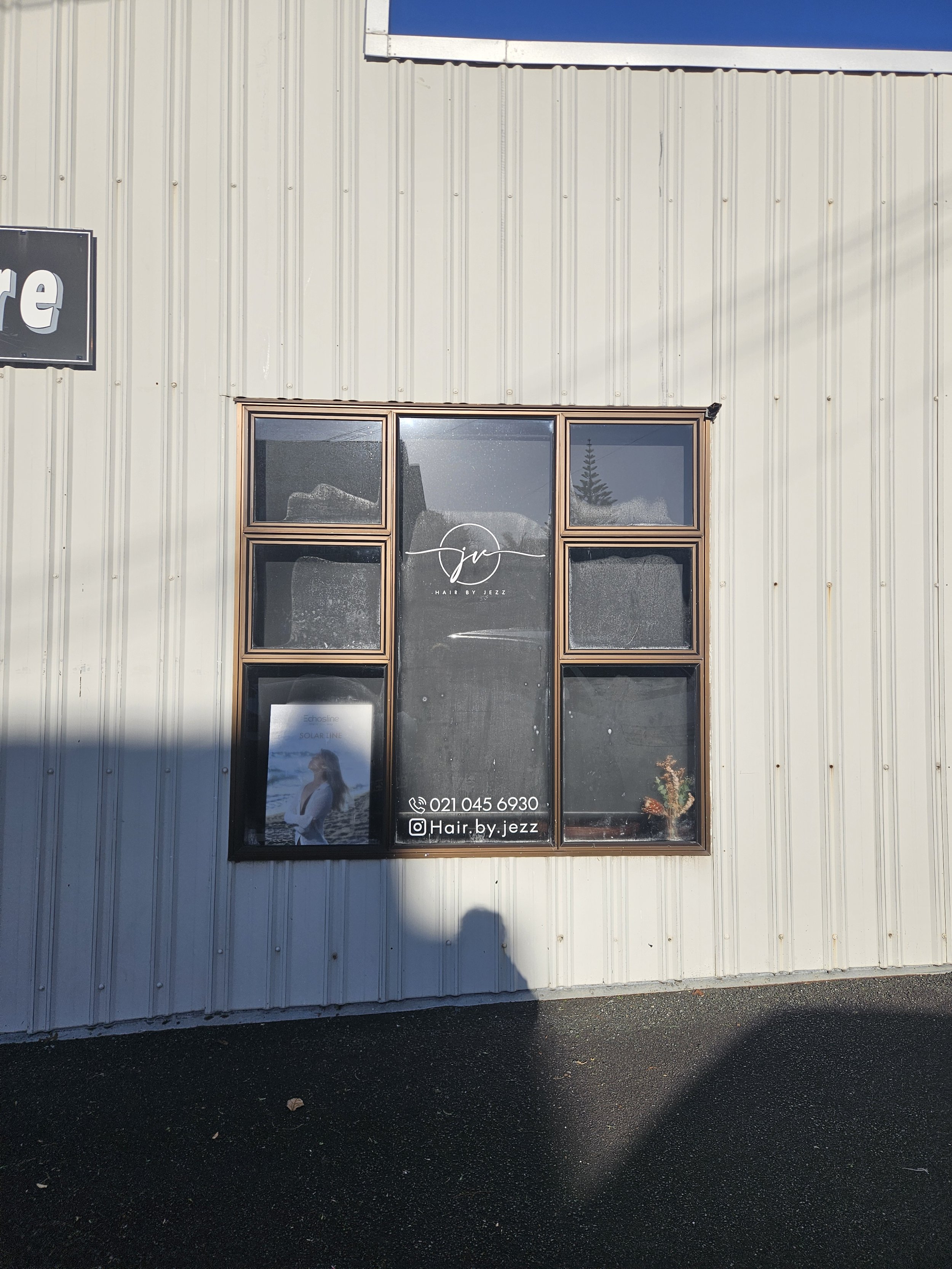 Photo of a building with corrugated metal exterior, large window with a sign for Hair by Jezz, and a poster of a woman with long hair at the bottom of the window.