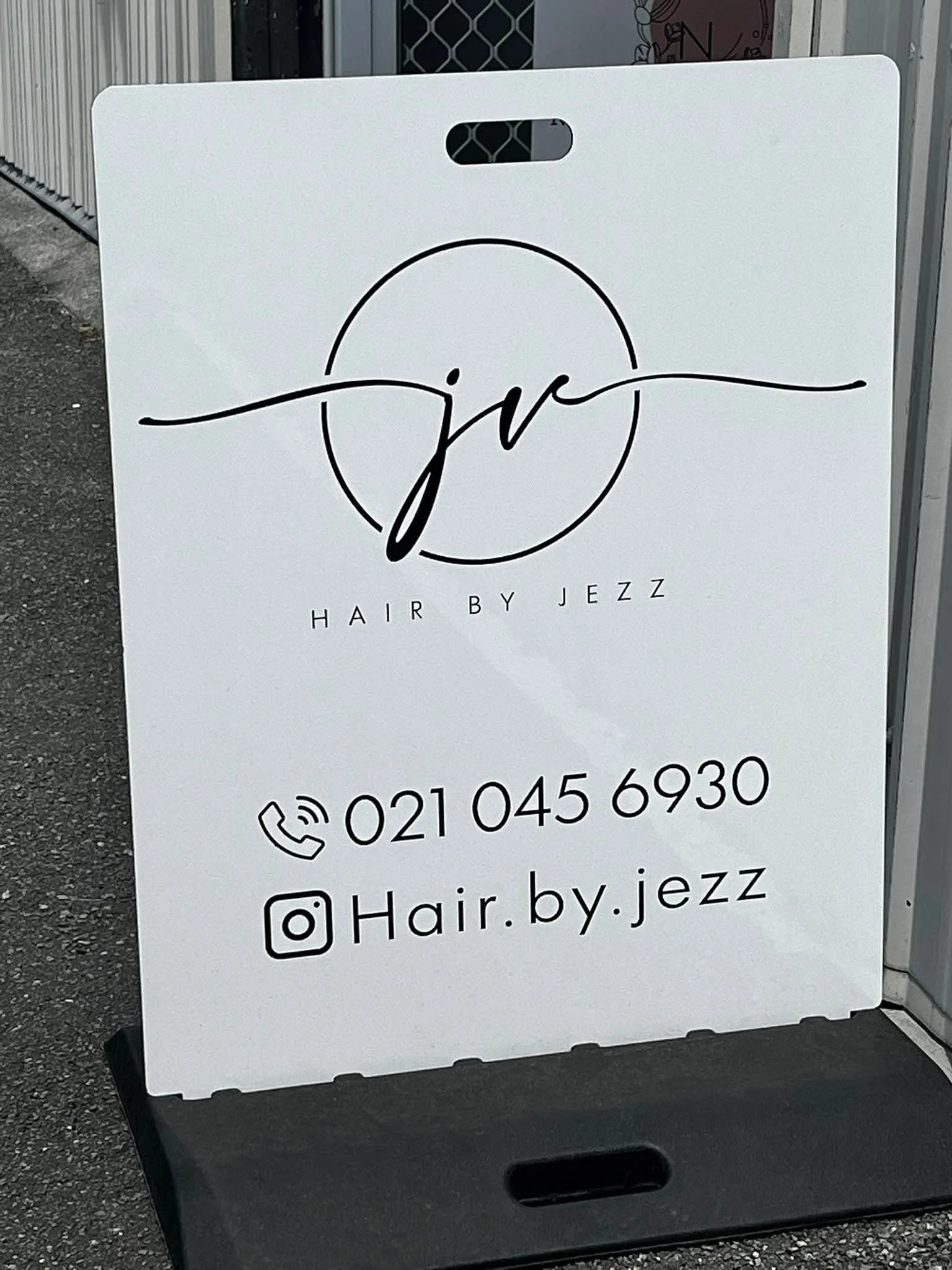 A white sidewalk sign for hair salon named Hair by Jezz with phone number and social media handles, placed on black base on pavement outside storefront.