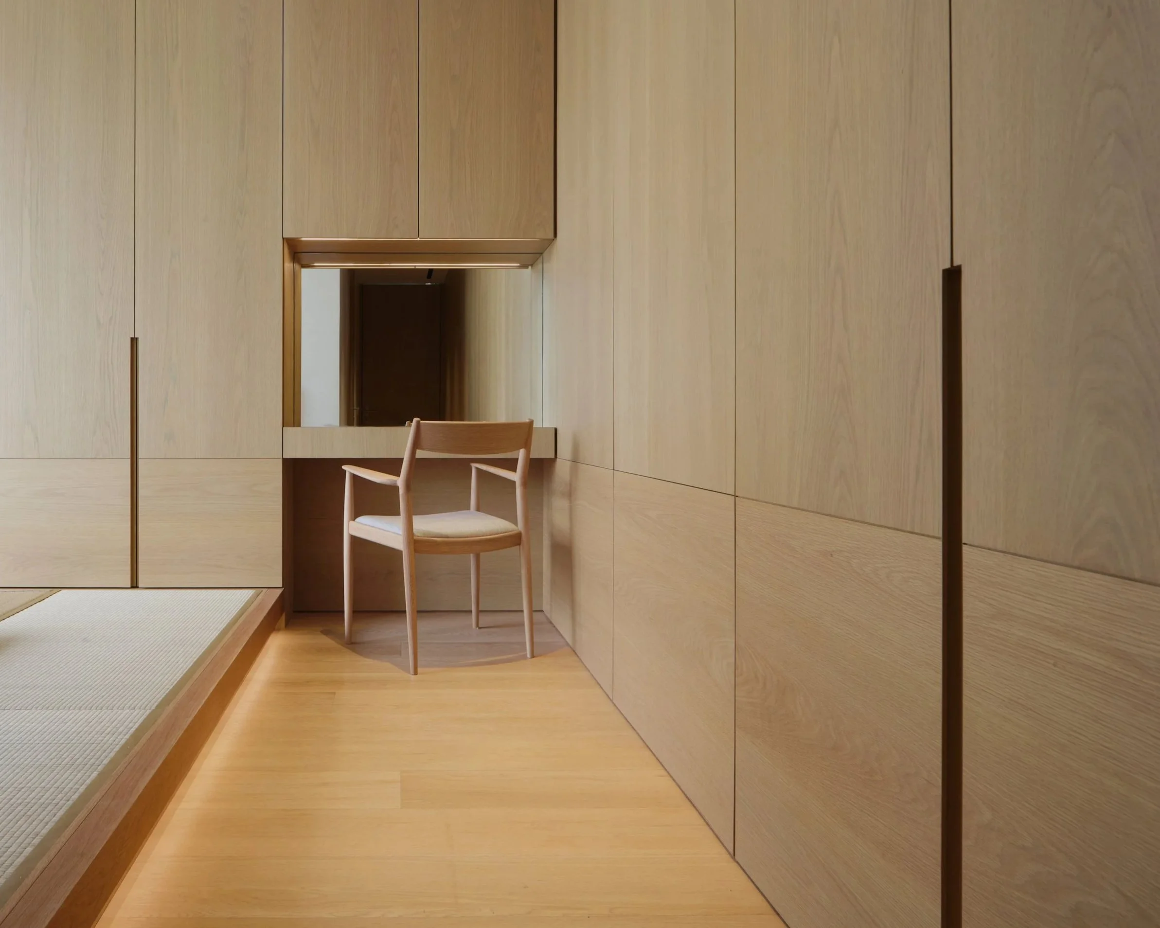 A room with wooden walls and floor, a telephone booth opening, a chair with a light wooden frame and beige cushioned seat, and a traditional Japanese tatami mat on the edge of the floor.