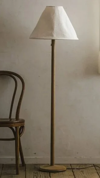 A floor lamp with a wooden pole and a beige lampshade next to a wooden chair against a plain wall.
