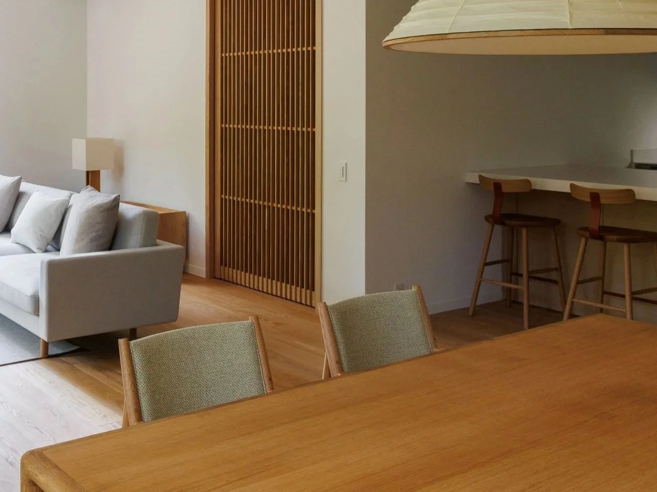 Living room with a beige sofa, wooden dining table, green upholstered chairs, a white bar counter with two wooden barstools, and a wooden sliding door.