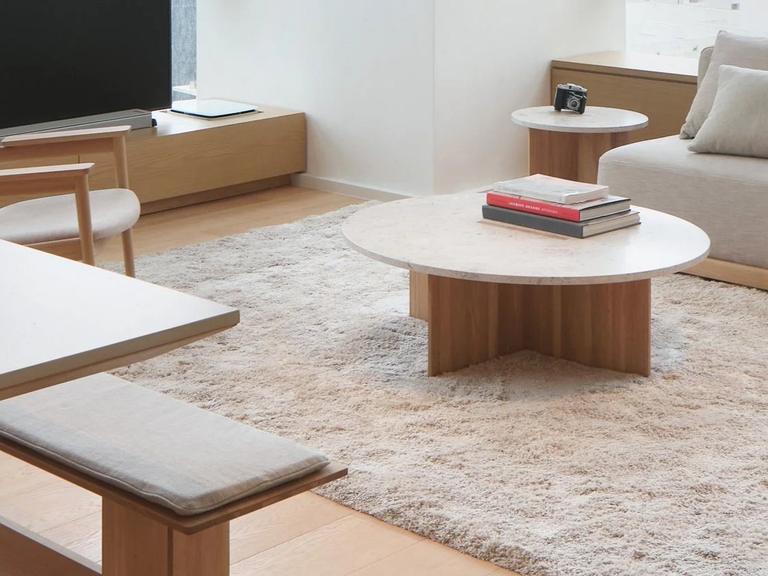 Modern living room with a white rug, a round marble-top coffee table with books, a beige sofa with pillows, a minimalist side table with a camera on it, a TV, and a wooden cabinet underneath the TV.