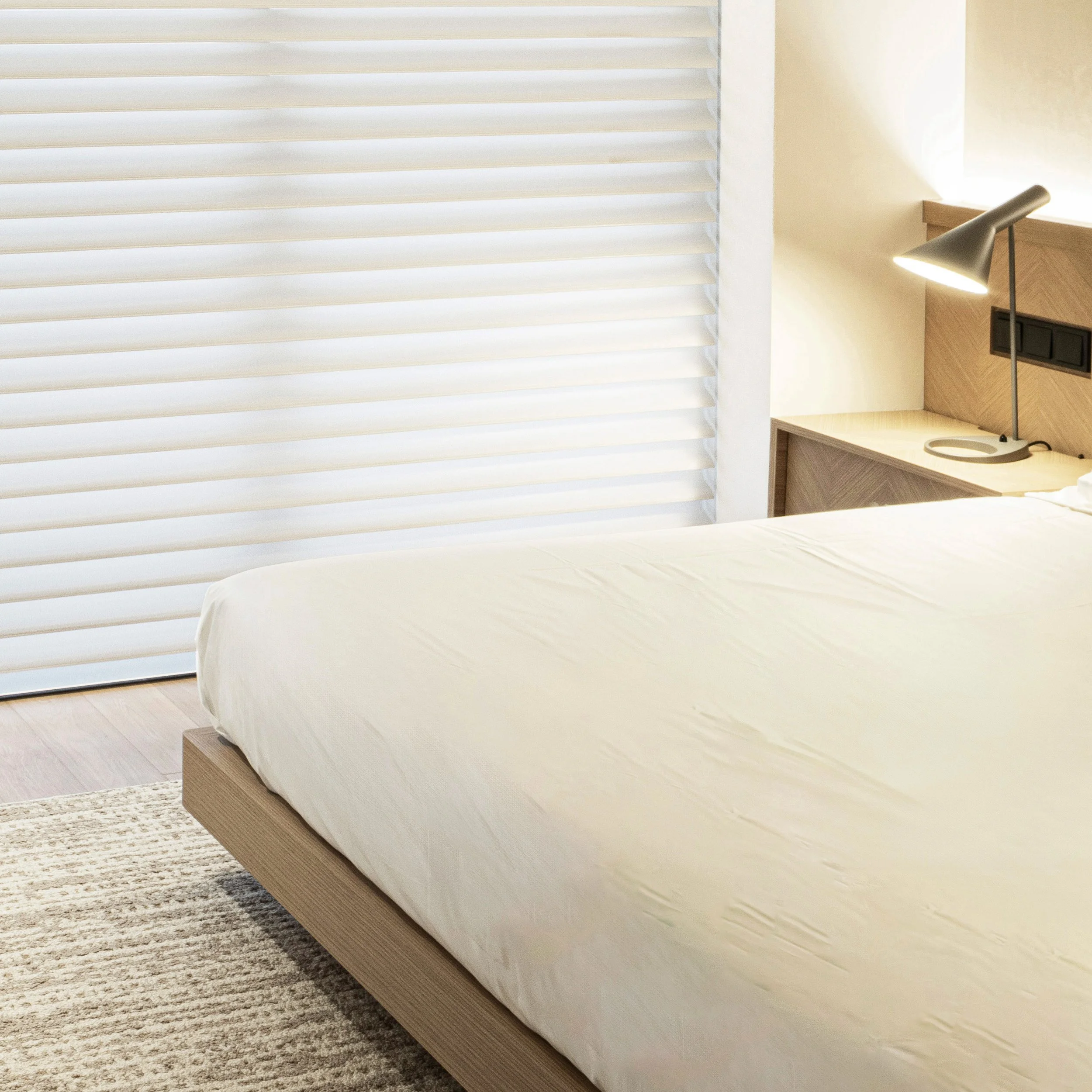 A bedroom with a bed covered in white sheets, a wooden headboard, a black metal desk lamp, a small desk or nightstand, and white window blinds covering a window.