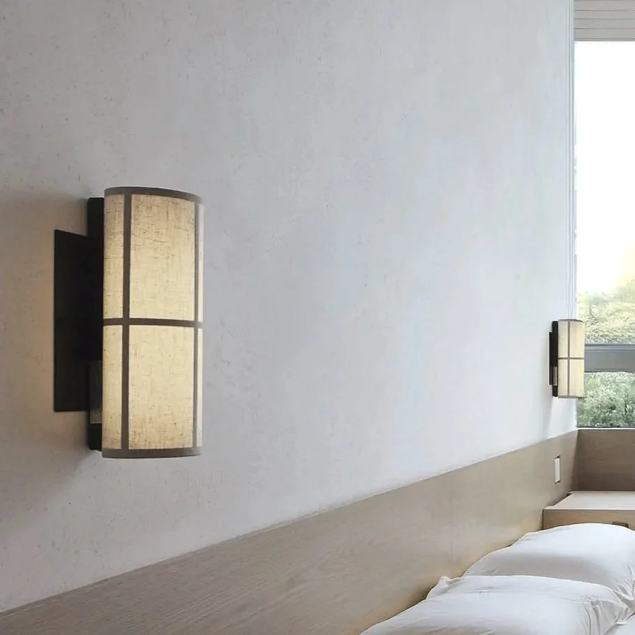 A bedroom with a beige headboard and two wall-mounted lamps with beige shades, one on each side of the bed. There is a window in the corner showing some greenery outside.