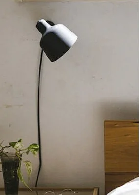 A white and black wall-mounted lamp over a bedside table with a potted plant, against a plain wall.